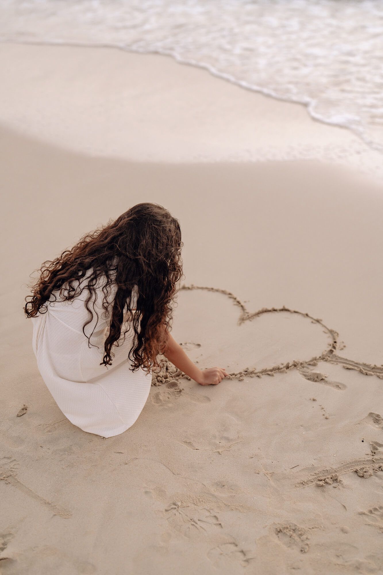 Person with long curly hair in white dress drawing heart shape in beach sand near ocean waves.