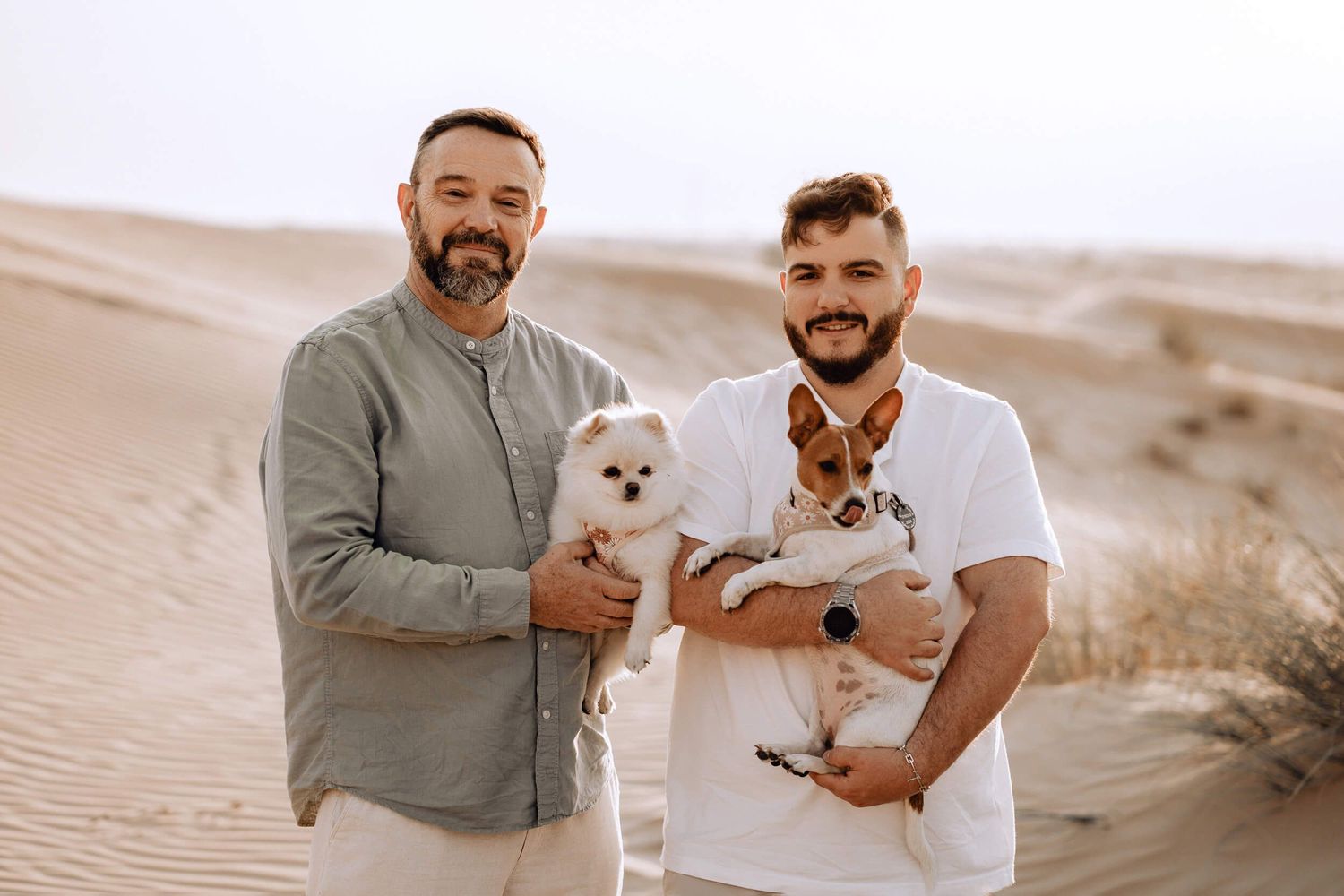 Two people pose with small white and brown dogs against desert sand dunes.