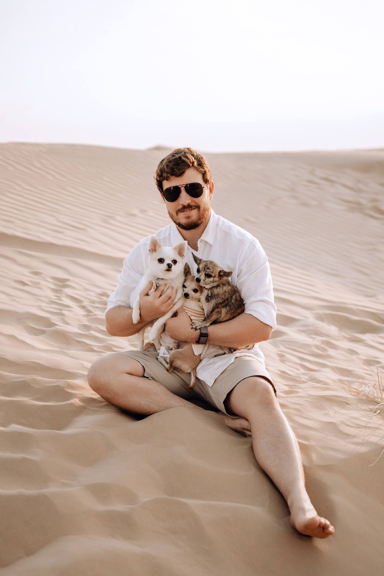Person sitting on desert sand holding small dog and cat while wearing sunglasses.