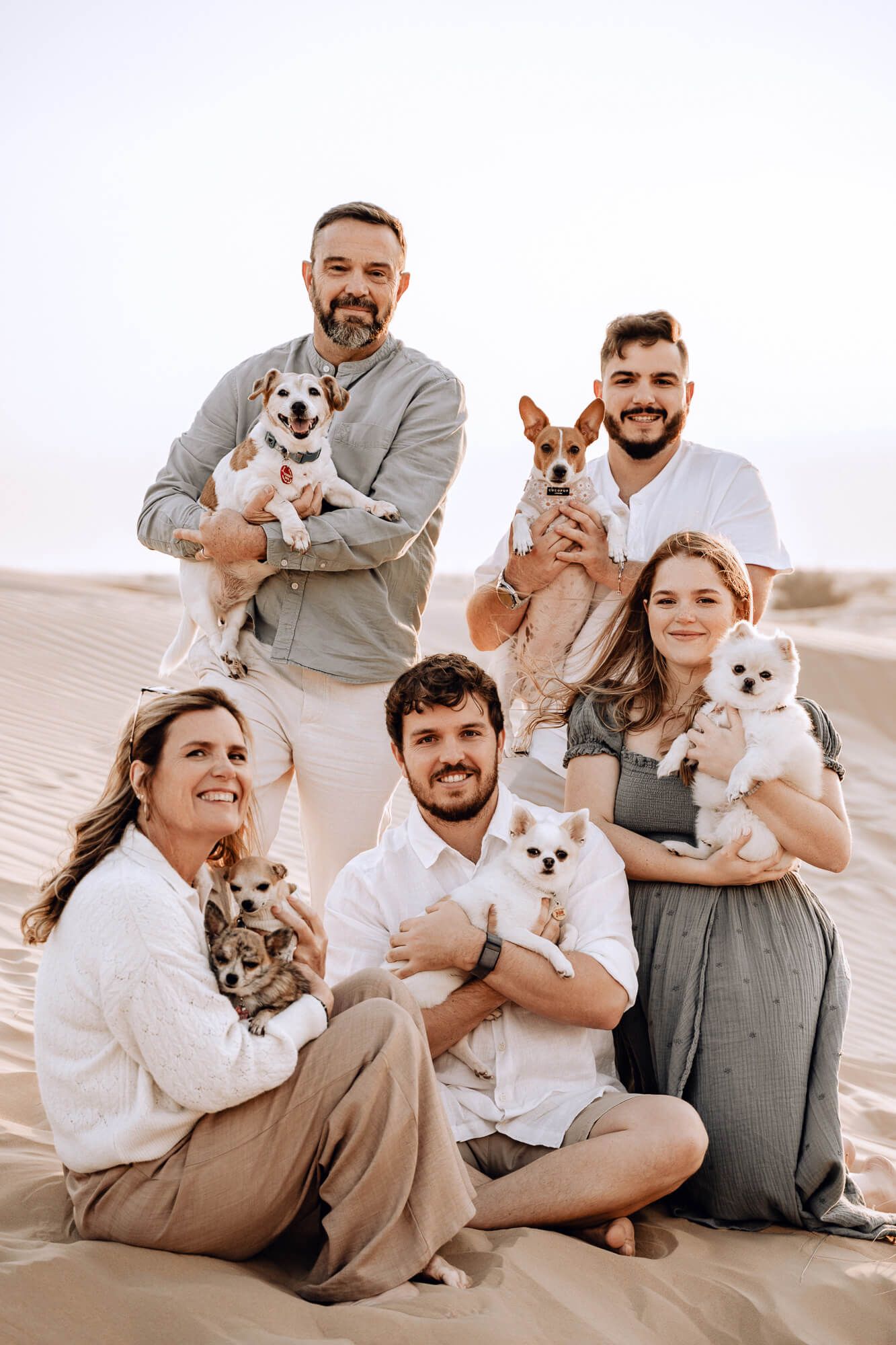 Desert photoshoot featuring five people holding their small dogs while sitting on sand dunes.