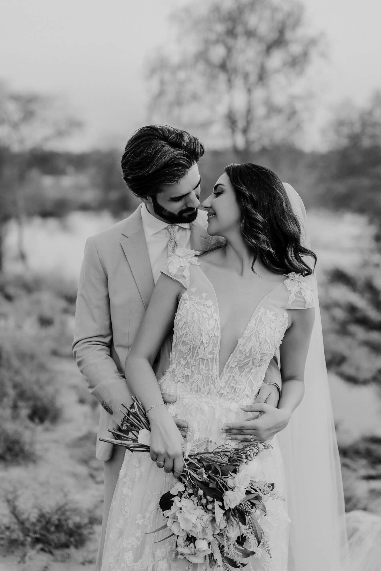 A romantic black and white wedding photo series of newlyweds embracing in a desert landscape with bouquet details.