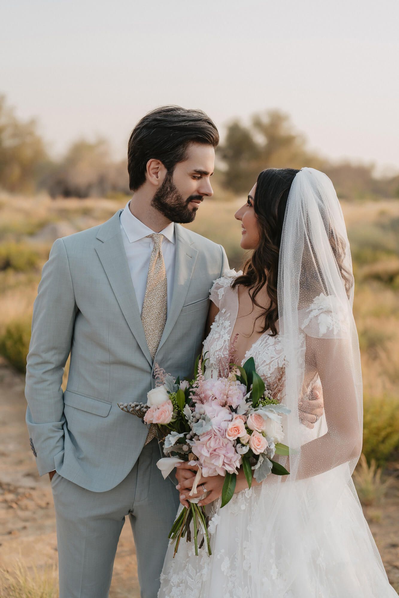Romantic desert wedding photos of couple in light gray suit and white lace gown with pink floral bouquet at sunset.