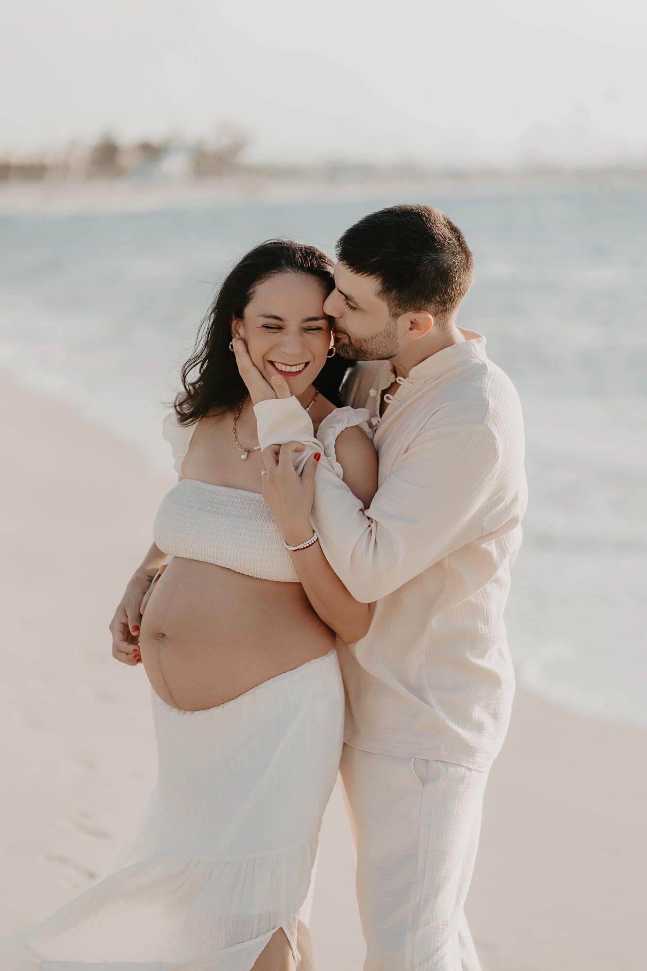 Romantic maternity photoshoot on a beach with couple in white outfits embracing at sunset.