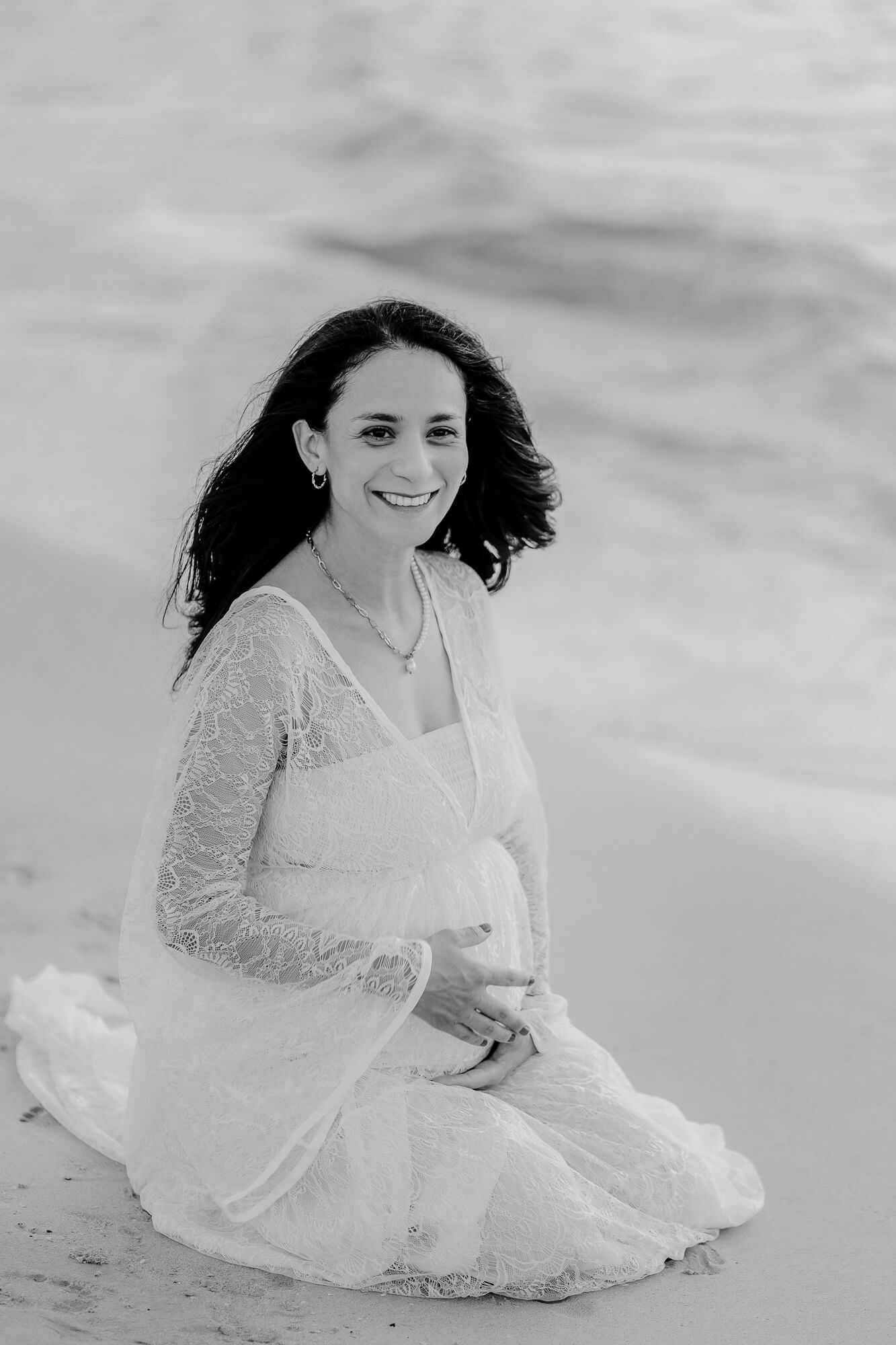 Black and white photo of someone sitting on a beach wearing a flowing white lace dress with windswept hair.