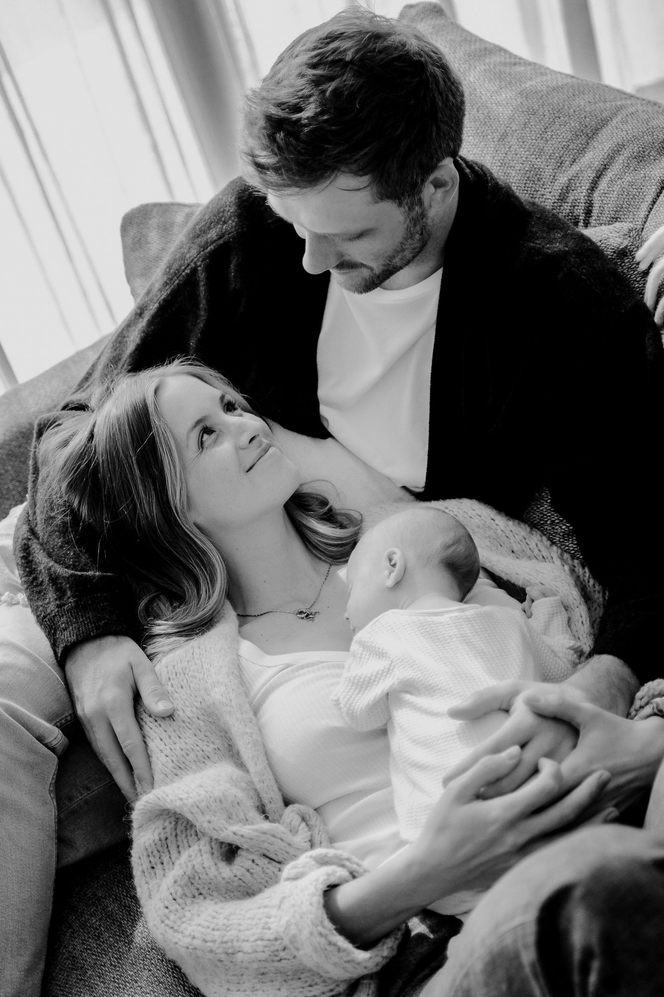 Black and white intimate family portrait showing parents lovingly gazing at their newborn baby while relaxing on a couch.