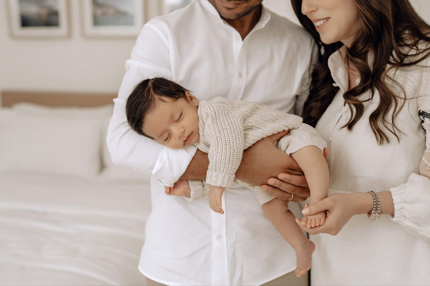 Intimate black and white family portrait with parents and sleeping baby on white bedding.