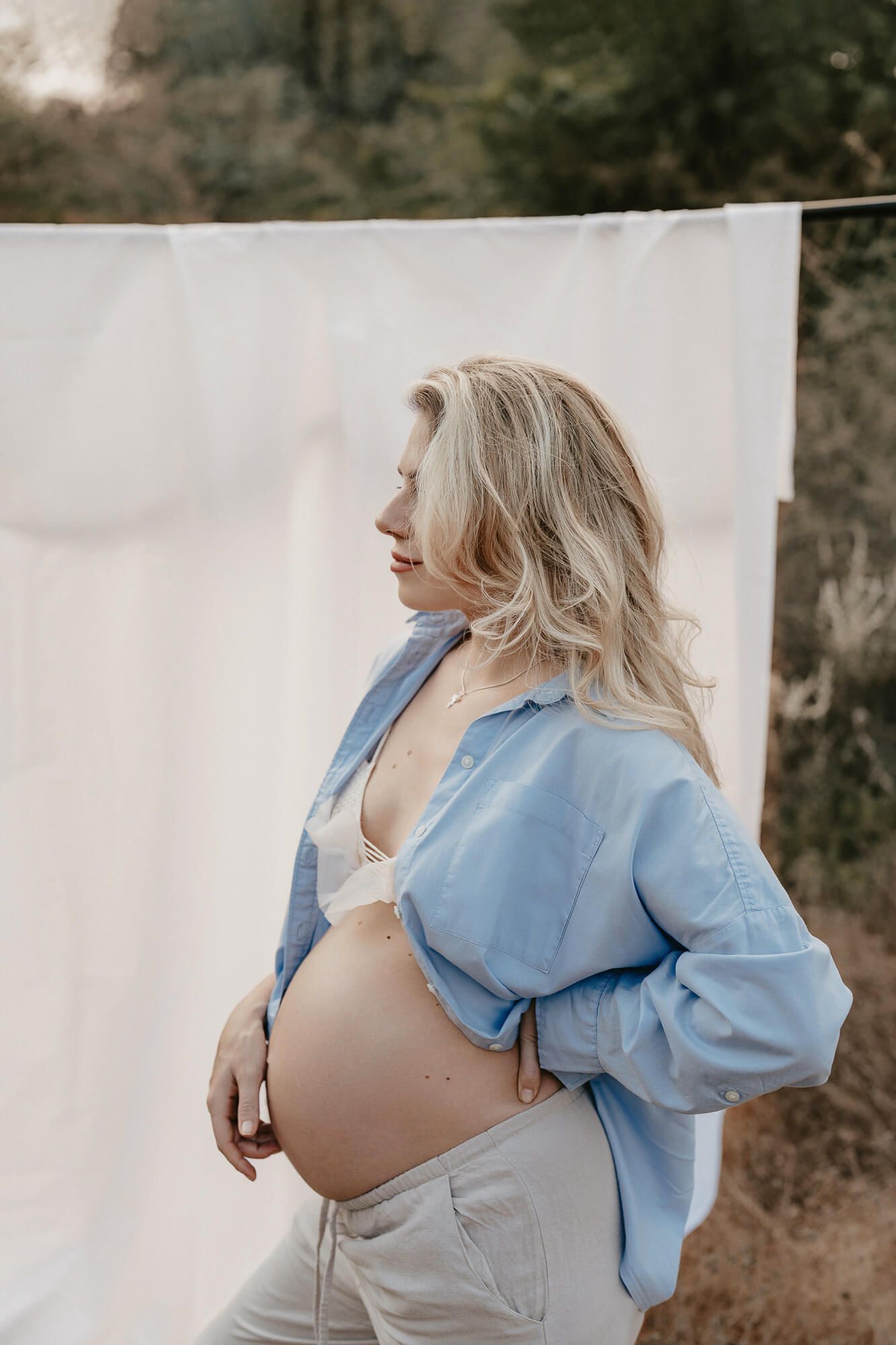 Side-view maternity portrait showcasing a blue shirt and casual style against white backdrop outdoors.