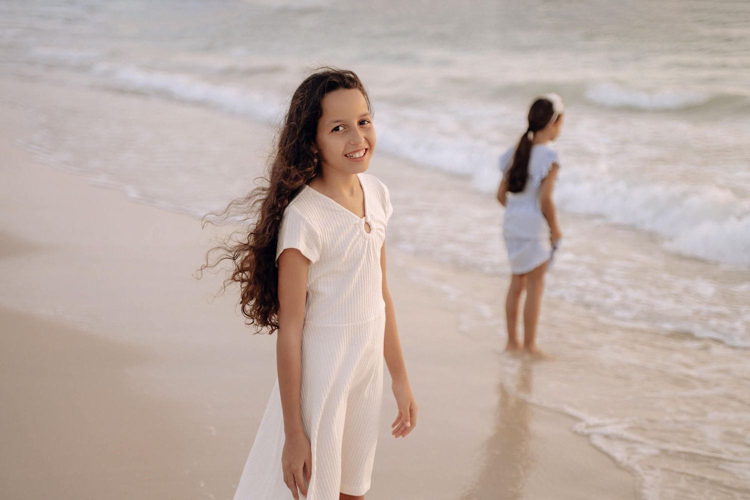 Two people in light dresses enjoy a peaceful moment walking along the shoreline.