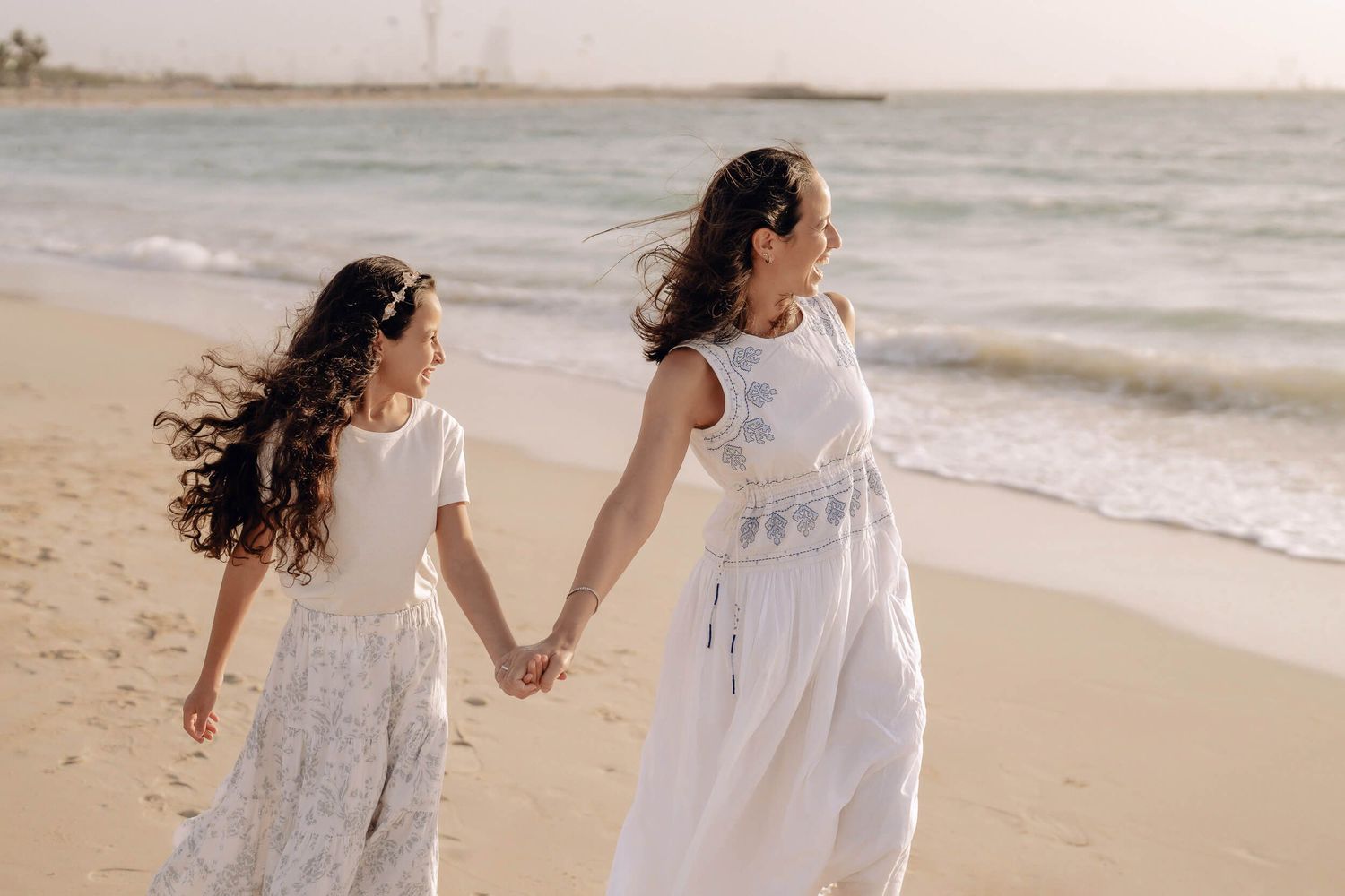 Two people in white dresses walk along the beach holding hands with wind blowing through their hair.