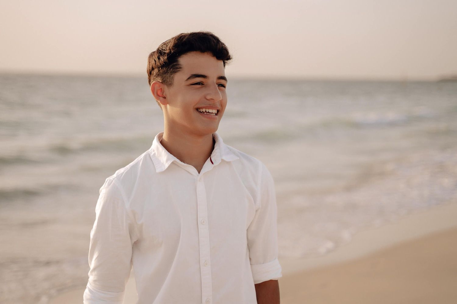 Young person smiling at sunset on a beach wearing a white button-down shirt.