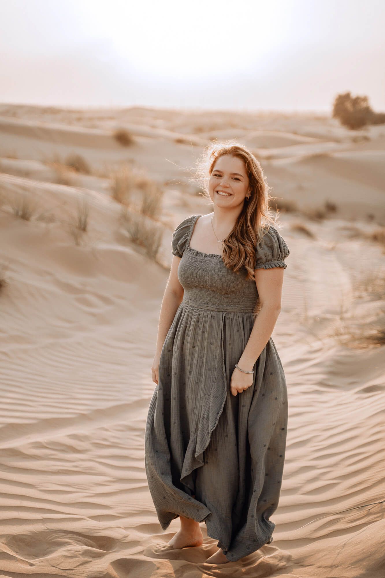 Woman in gray dress poses elegantly on desert sand dunes during sunset.