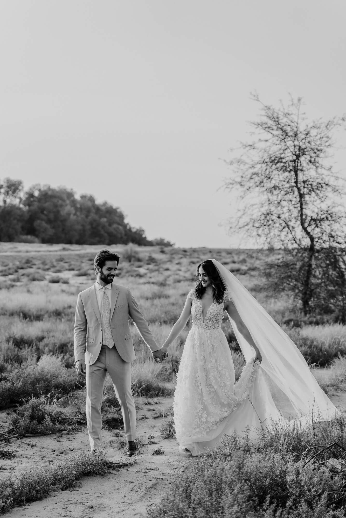 A romantic black and white desert wedding photo of a couple holding hands on a path with flowing veil and desert brush.