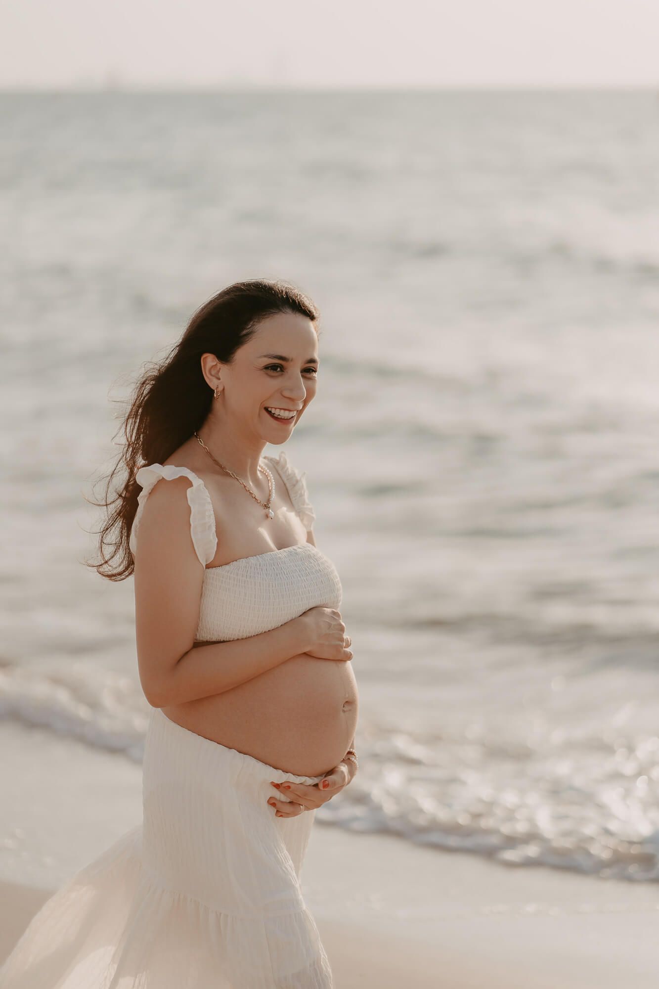 A pregnant woman in a white dress walks along a serene beach during golden hour, cradling her baby bump.