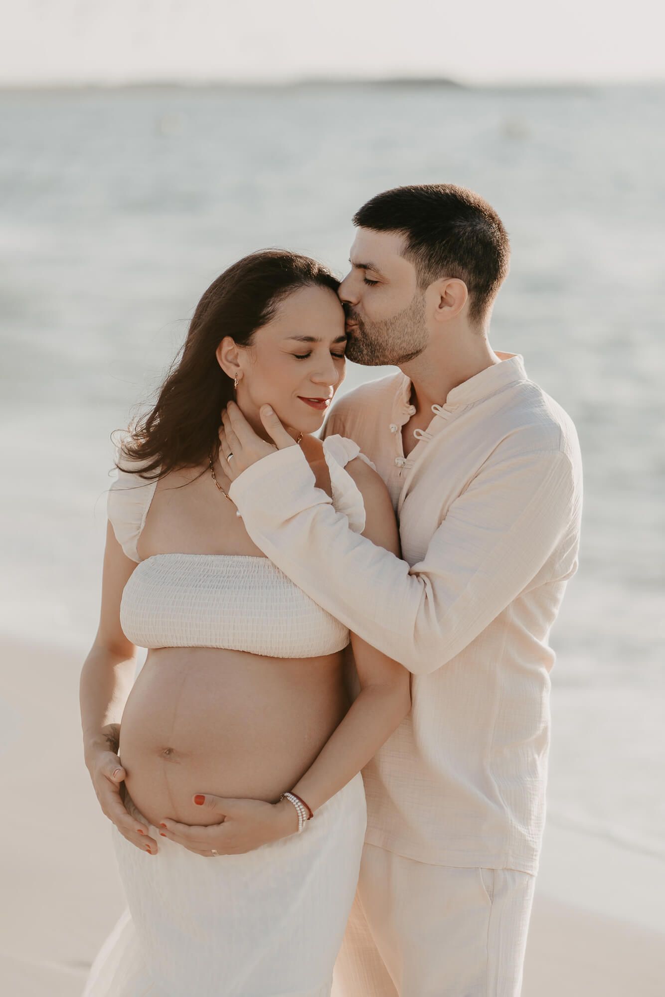 Intimate maternity beach photo of a couple in white clothing embracing as waves roll in the background.