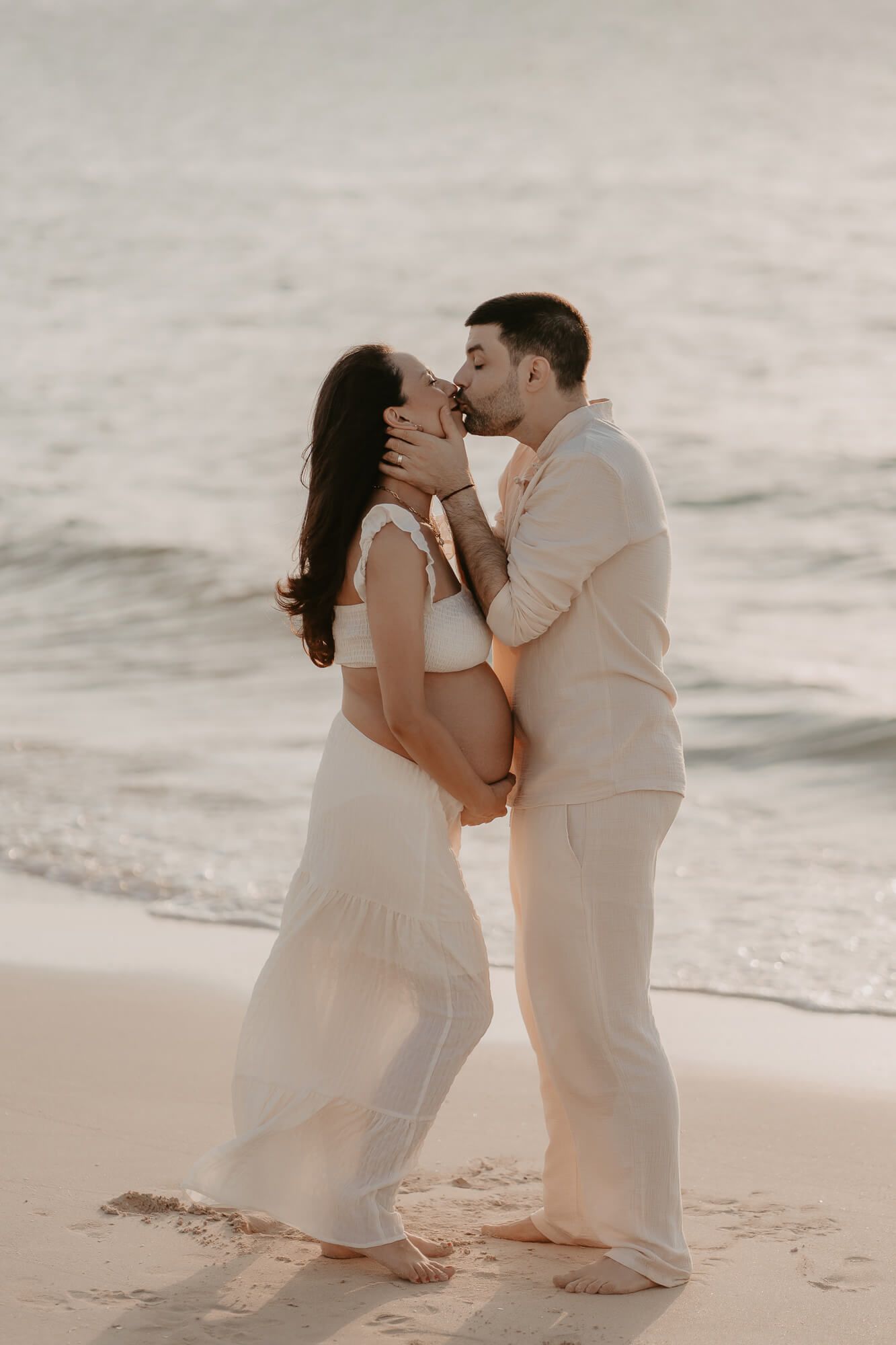 Romantic couple sharing an intimate moment in white attire on a peaceful beach at sunset.