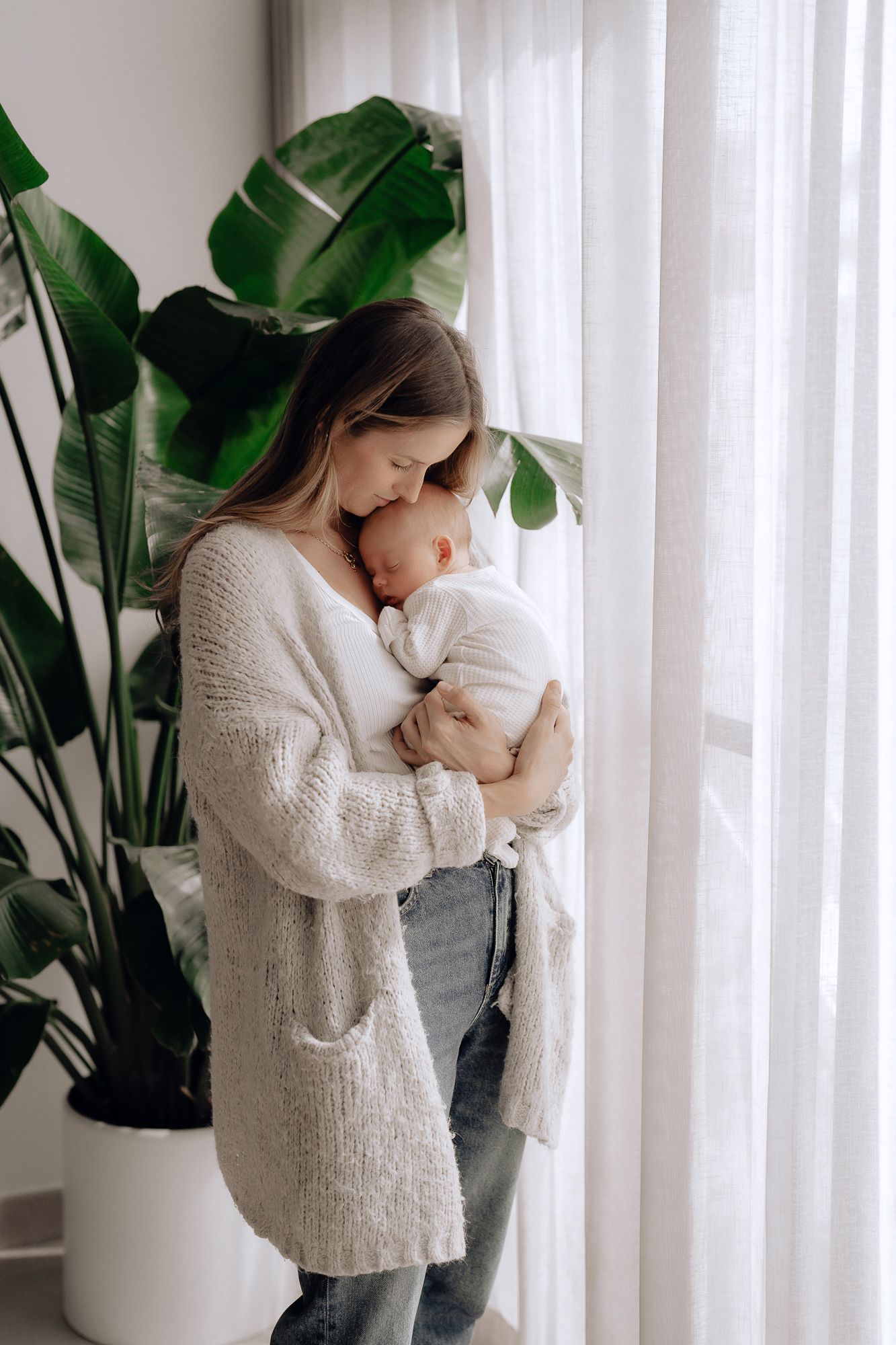 A gentle embrace between parent and child by a window with a monstera plant in the background.