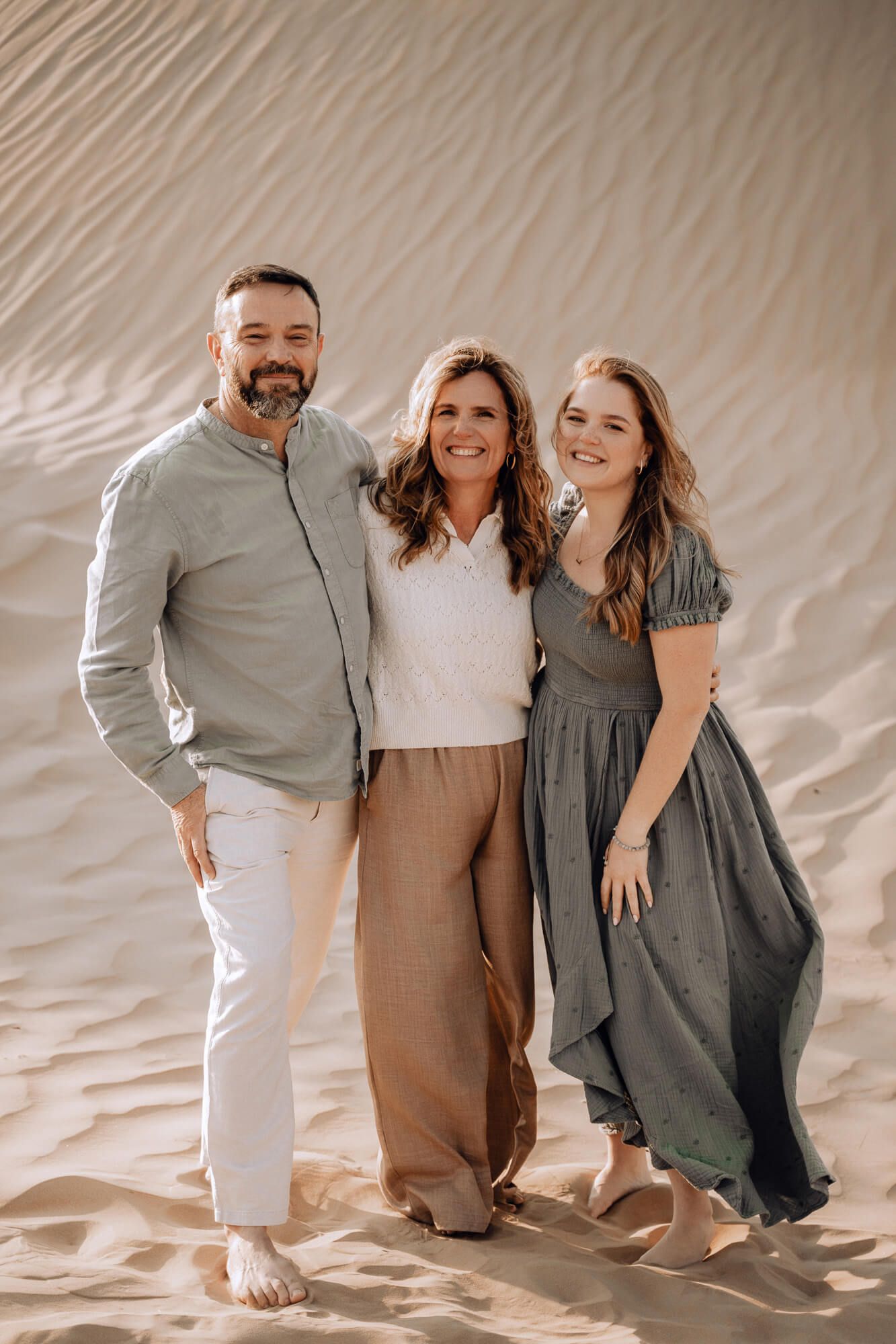 Family portrait on sand dunes with three people wearing coordinated neutral-toned outfits.