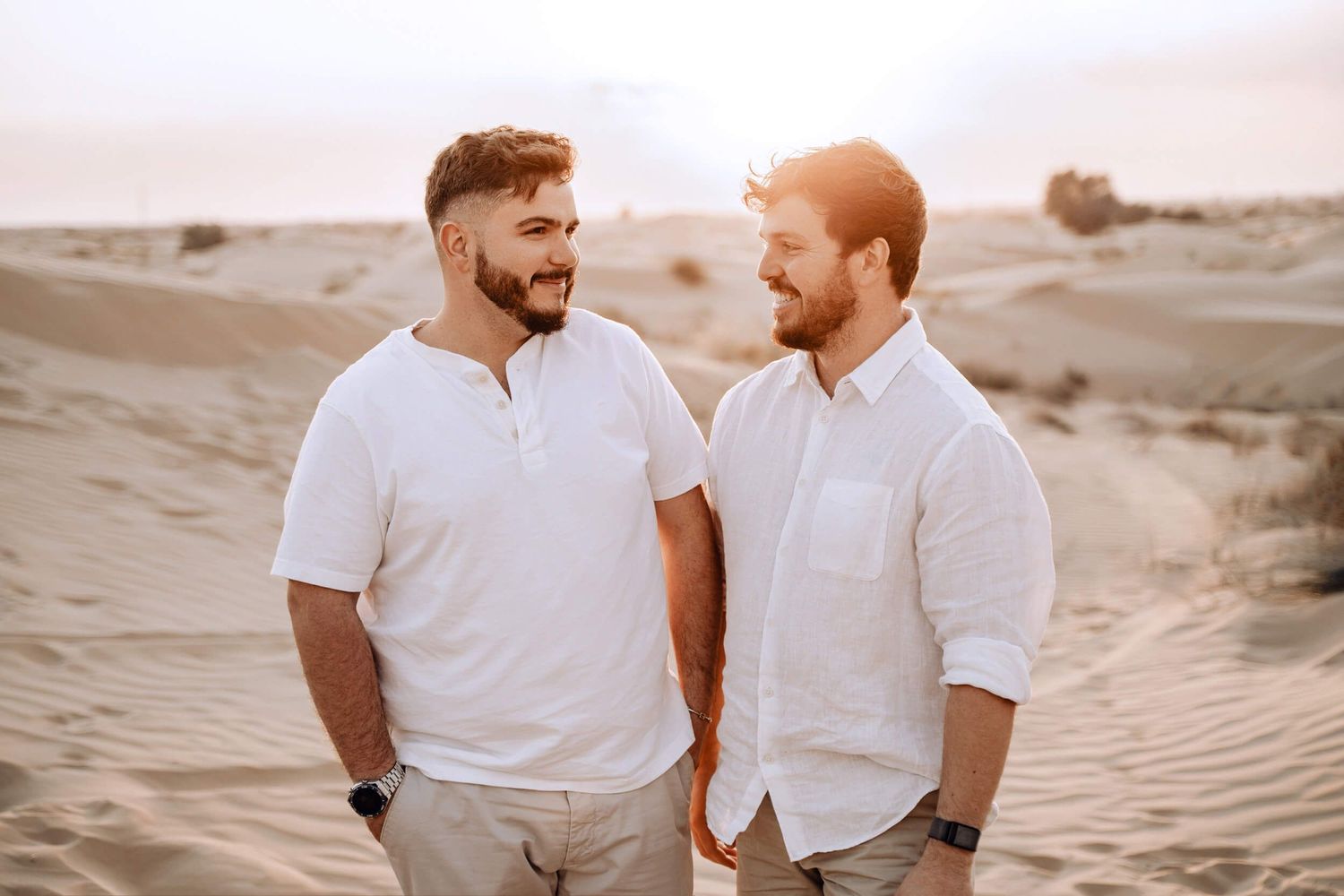 Two people in white shirts share a candid moment while standing in desert sand dunes.