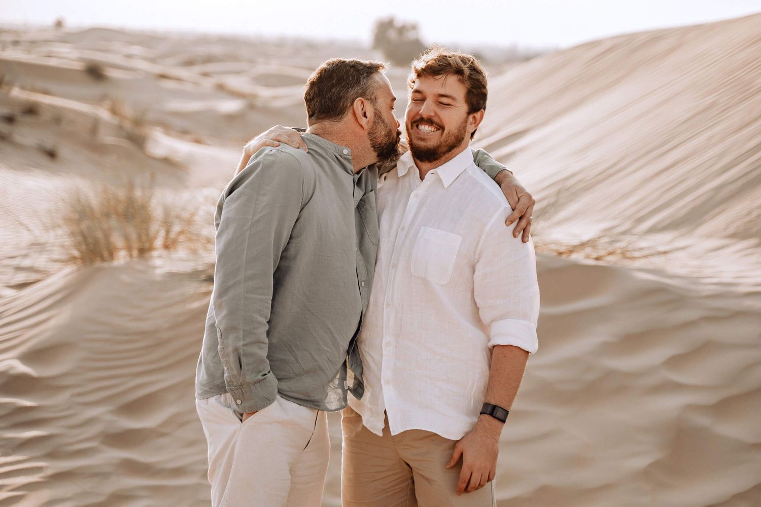 Two people share a loving embrace against sandy desert dunes in warm sepia tones.
