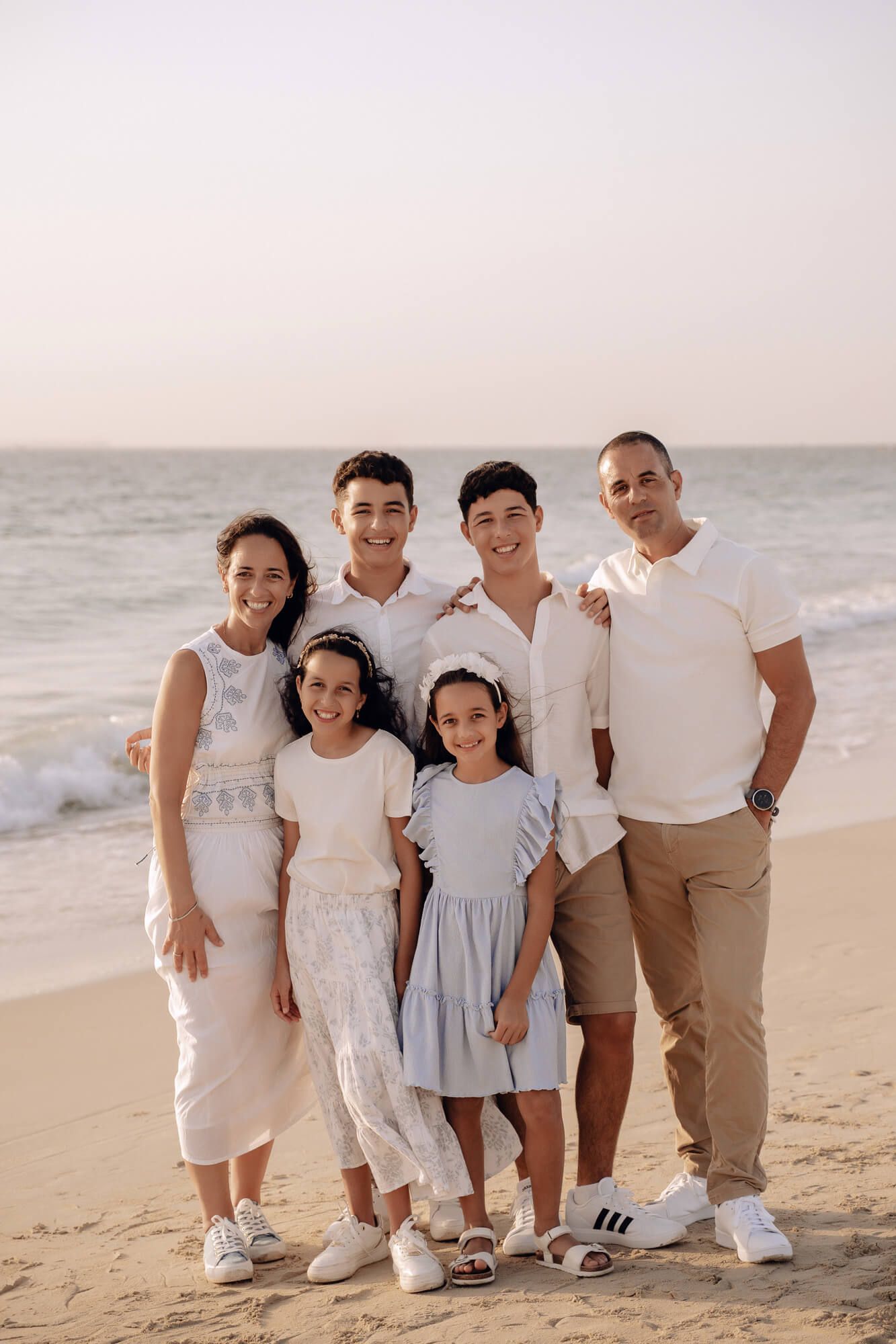 Group portrait of six people wearing matching white and neutral outfits on a sandy beach.