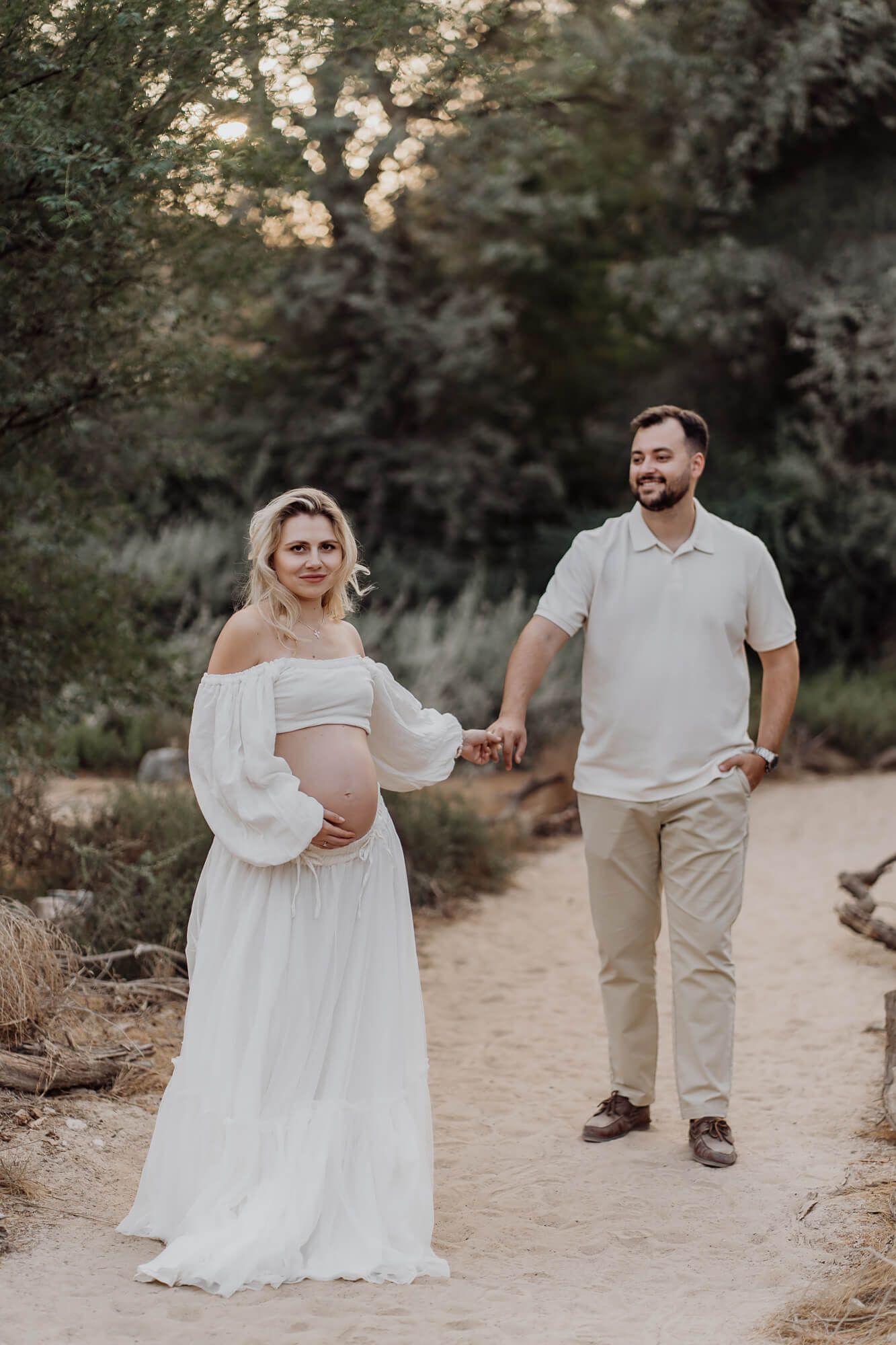 Elegant maternity session photographed on a sandy path surrounded by desert vegetation.