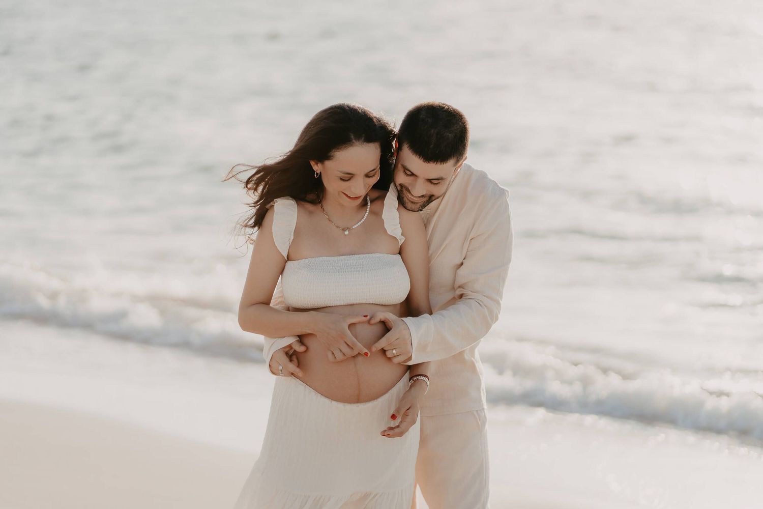 A couple in white and beige outfits share an intimate moment on a beach during a maternity photoshoot.