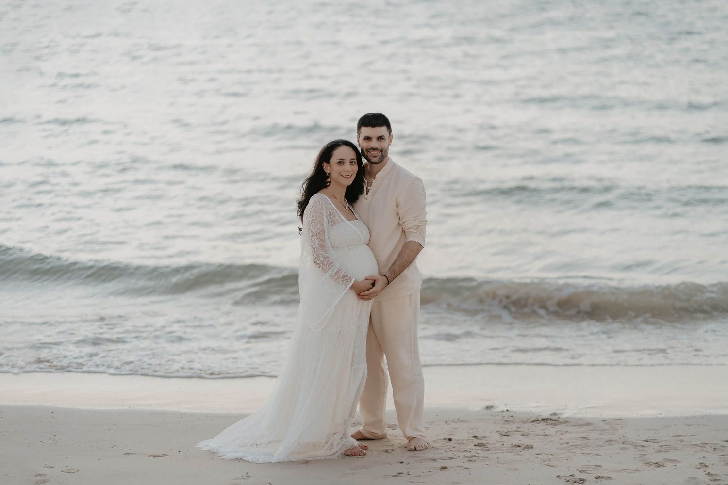 A couple in white attire embraces on a serene beach with ocean waves in the background during sunset.