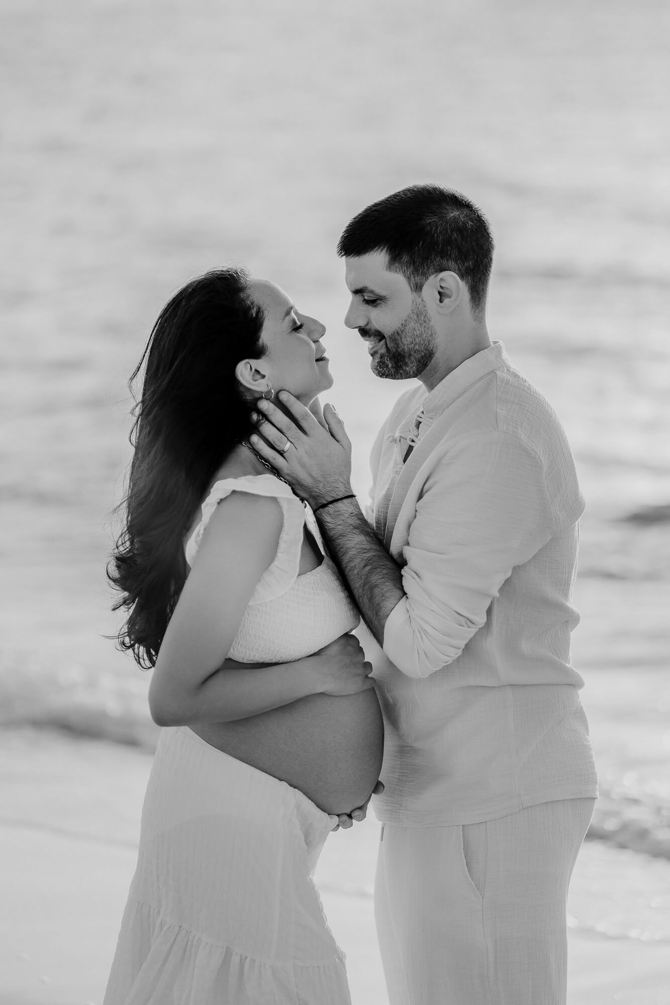 Romantic black and white maternity photo of a couple in white attire embracing on a beach.