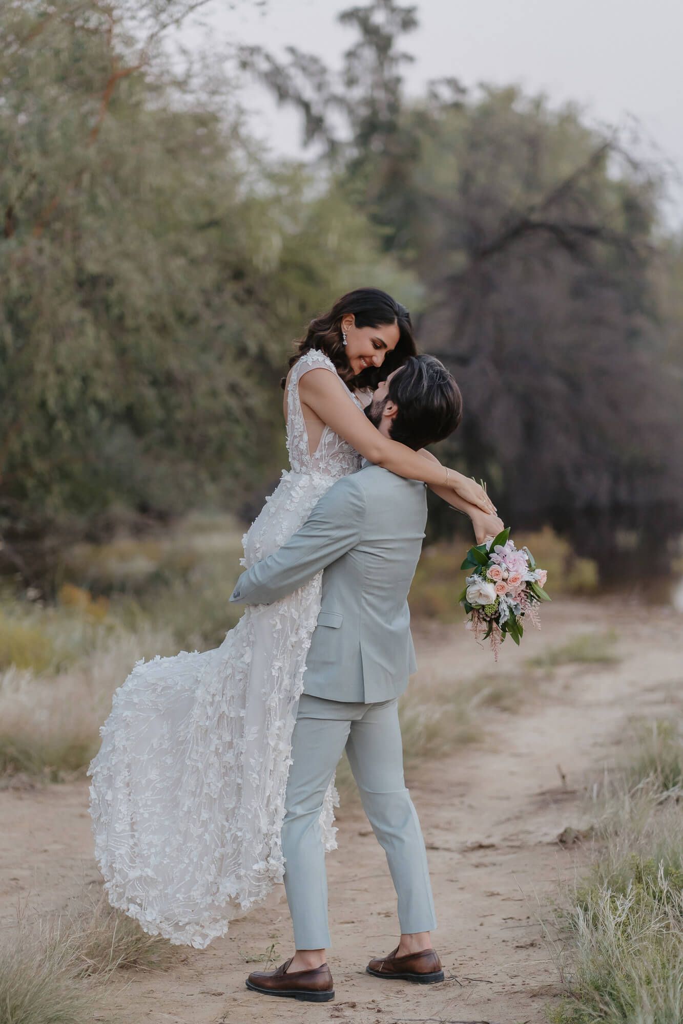 A romantic couple in light blue suit and lace dress share tender moments during their desert wedding photo session.