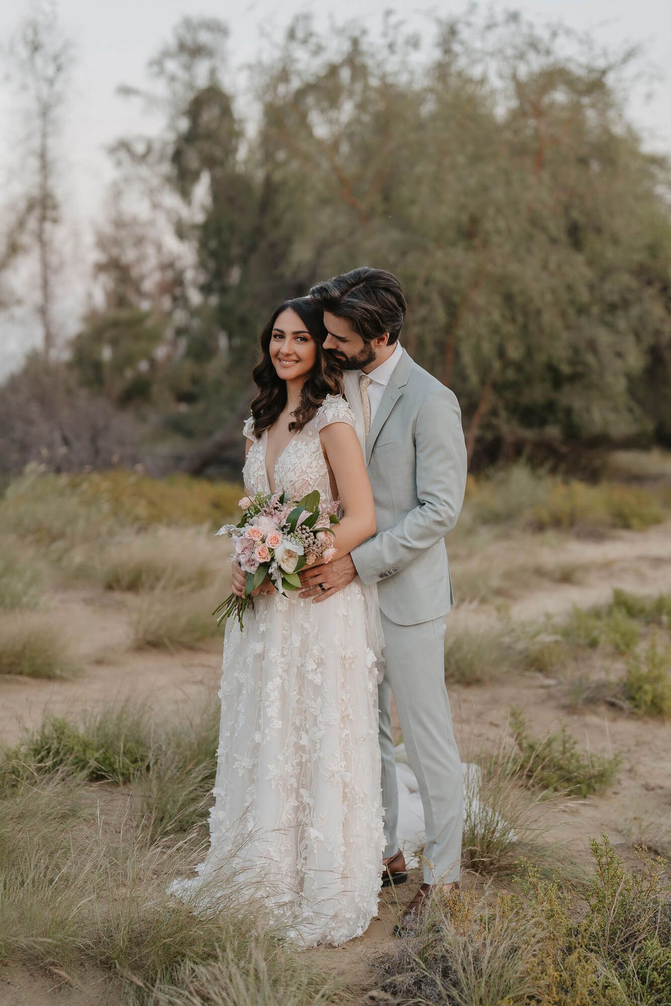 A romantic wedding couple in grey suit and lace dress embrace in a field of tall grass at sunset.