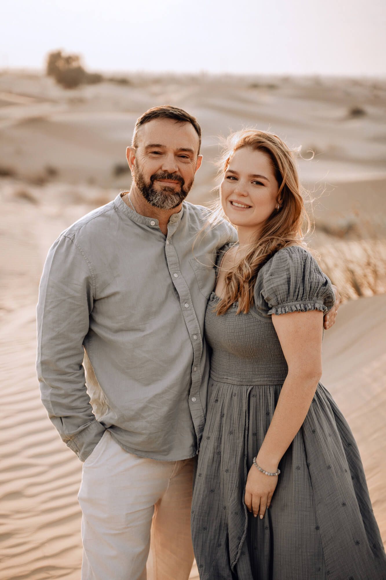 A couple poses together on desert sand dunes wearing coordinated gray and beige outfits in warm lighting.