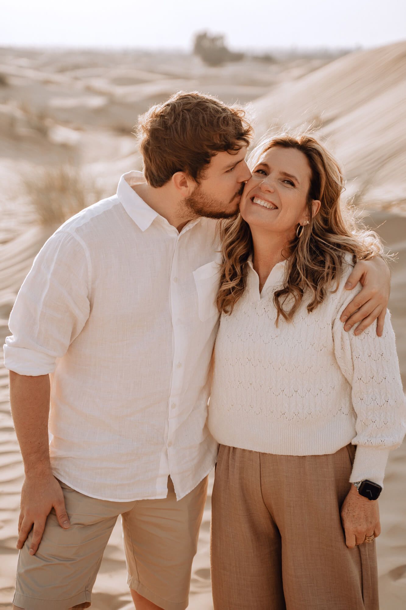 Tender desert photoshoot moment with neutral earth-toned clothing against sandy dunes.