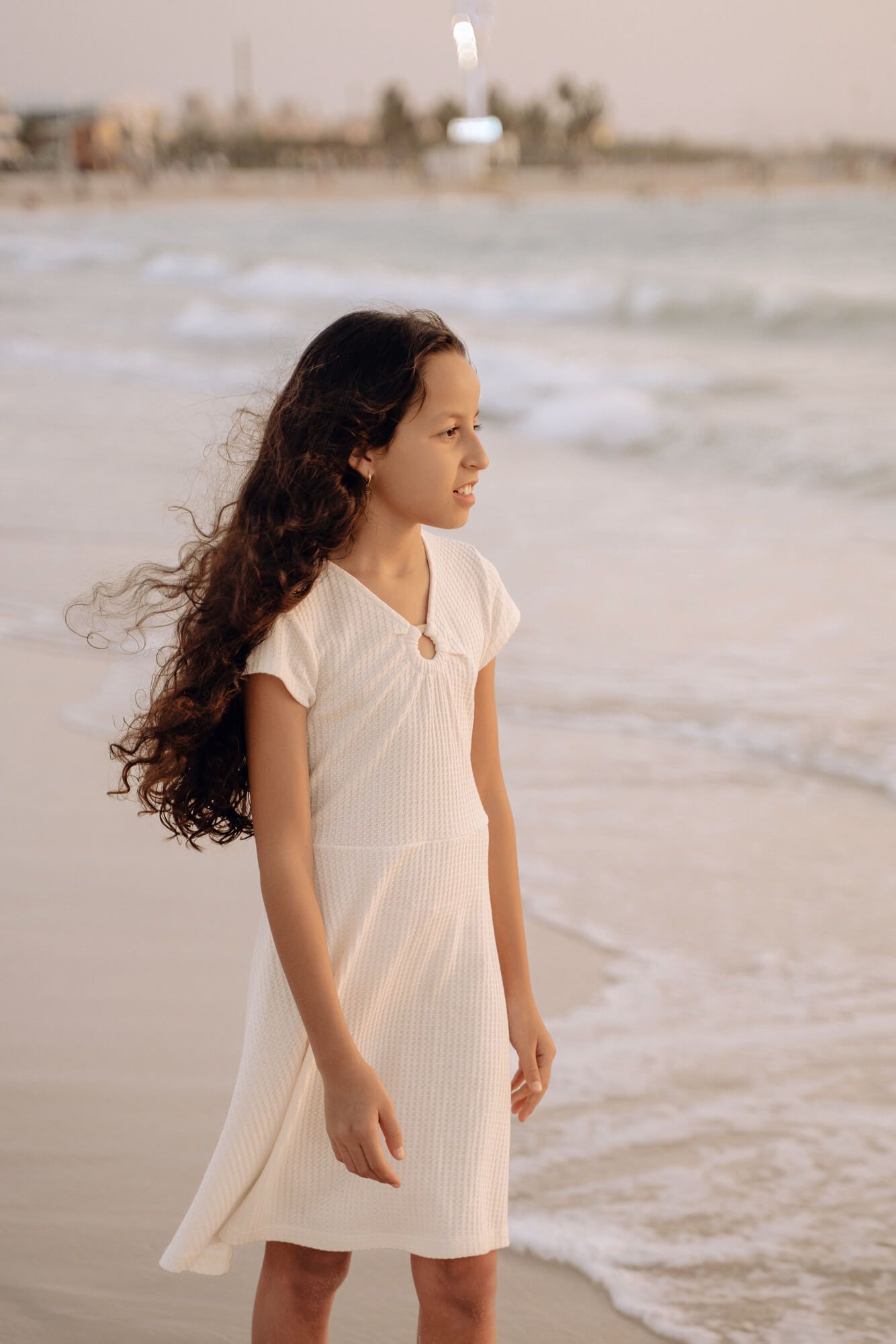 A person in a white ribbed dress stands on the beach with hair flowing in the wind.
