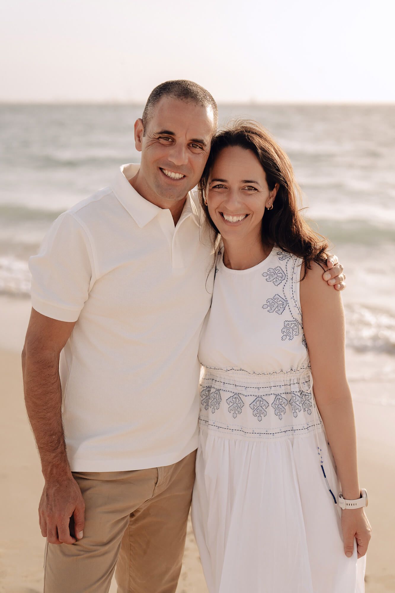 A couple wearing coordinated white outfits smile together on a beach with ocean waves behind them.