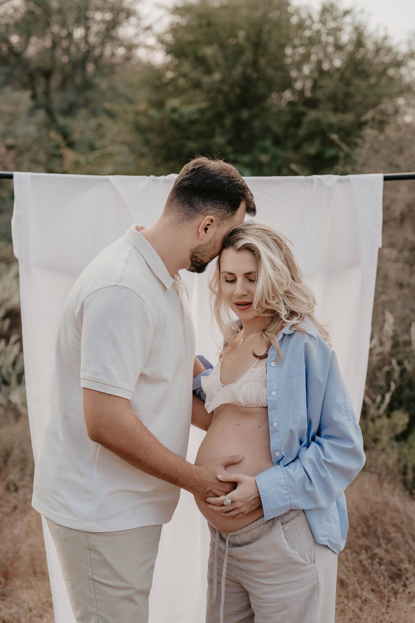 Intimate maternity photoshoot featuring a couple in casual wear against a white backdrop outdoors.