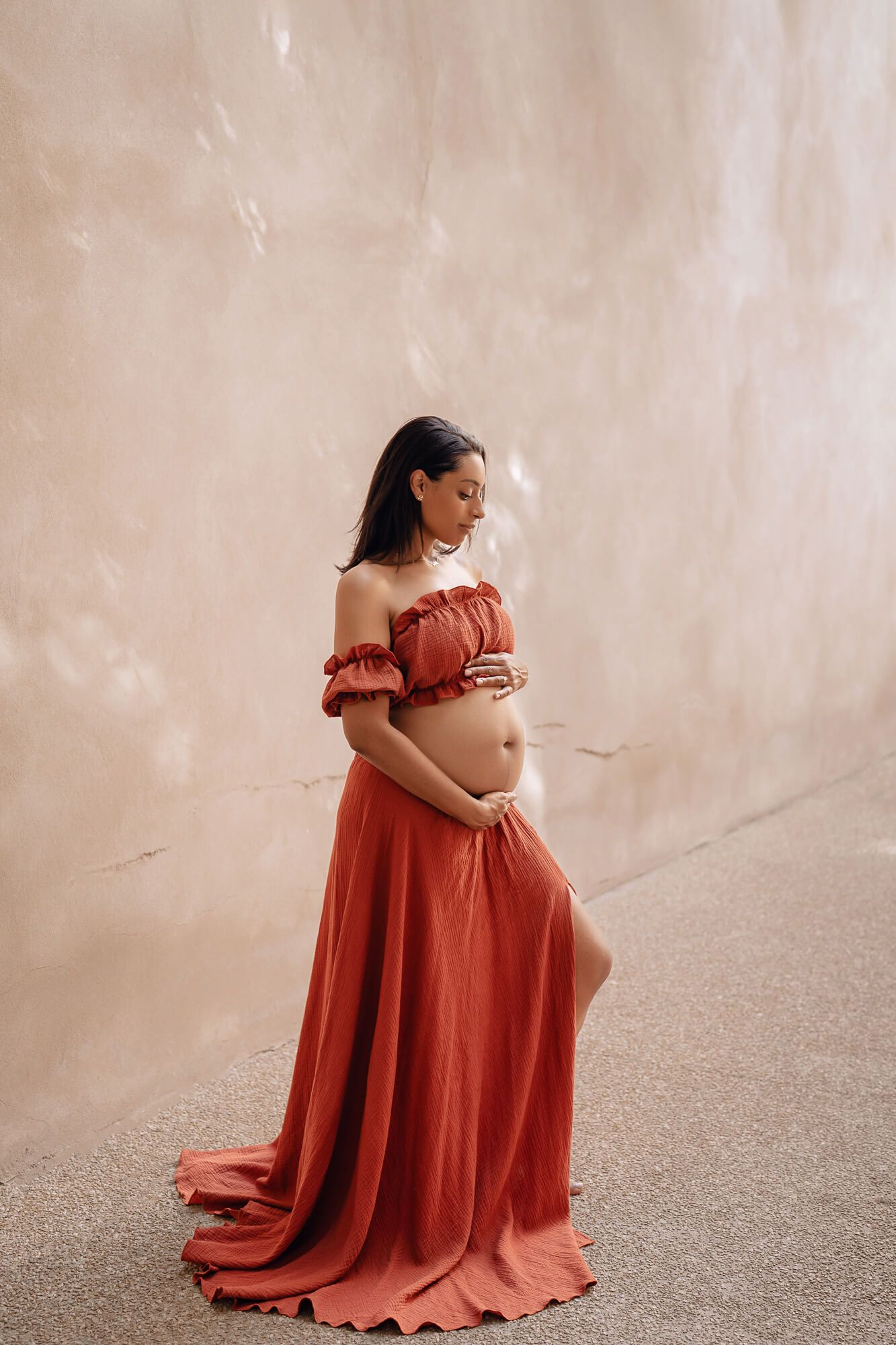 Expectant mother in ruffled terracotta dress posing gracefully on concrete floor.
