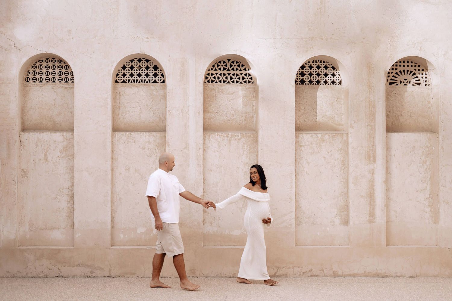 A couple in white outfits holds hands against an architectural backdrop with arched alcoves.