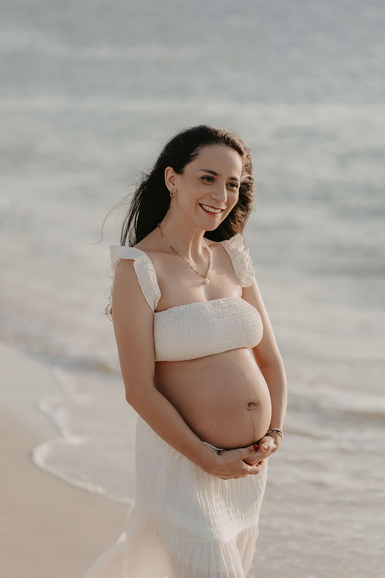 Expecting mother in flowing white dress lovingly cradles baby bump during sunset maternity photoshoot at beach.