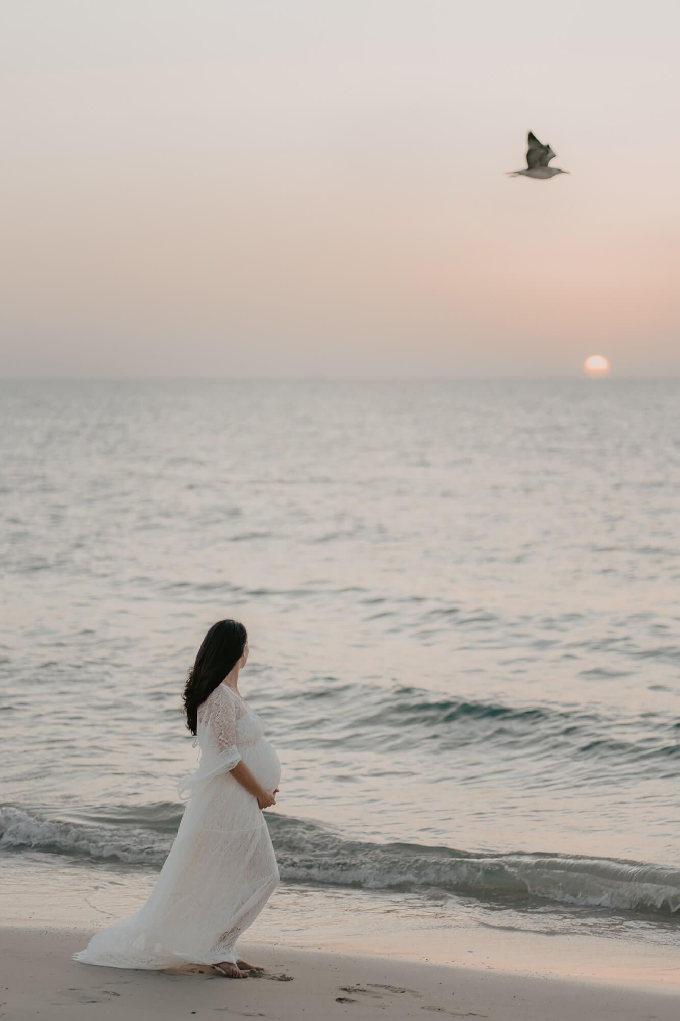 A dreamy coastal scene shows an expectant moth in a flowing white dress sitting on the beach at sunset watching seabirds fly over the ocean.