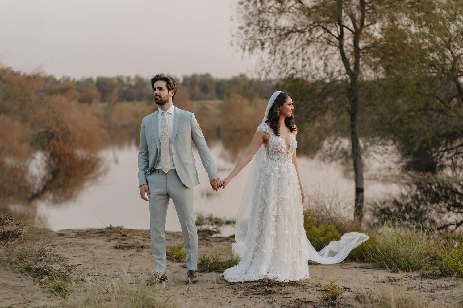 A bride and groom hold hands while standing by a tranquil lake during autumn, with the bride's dress flowing in the wind.