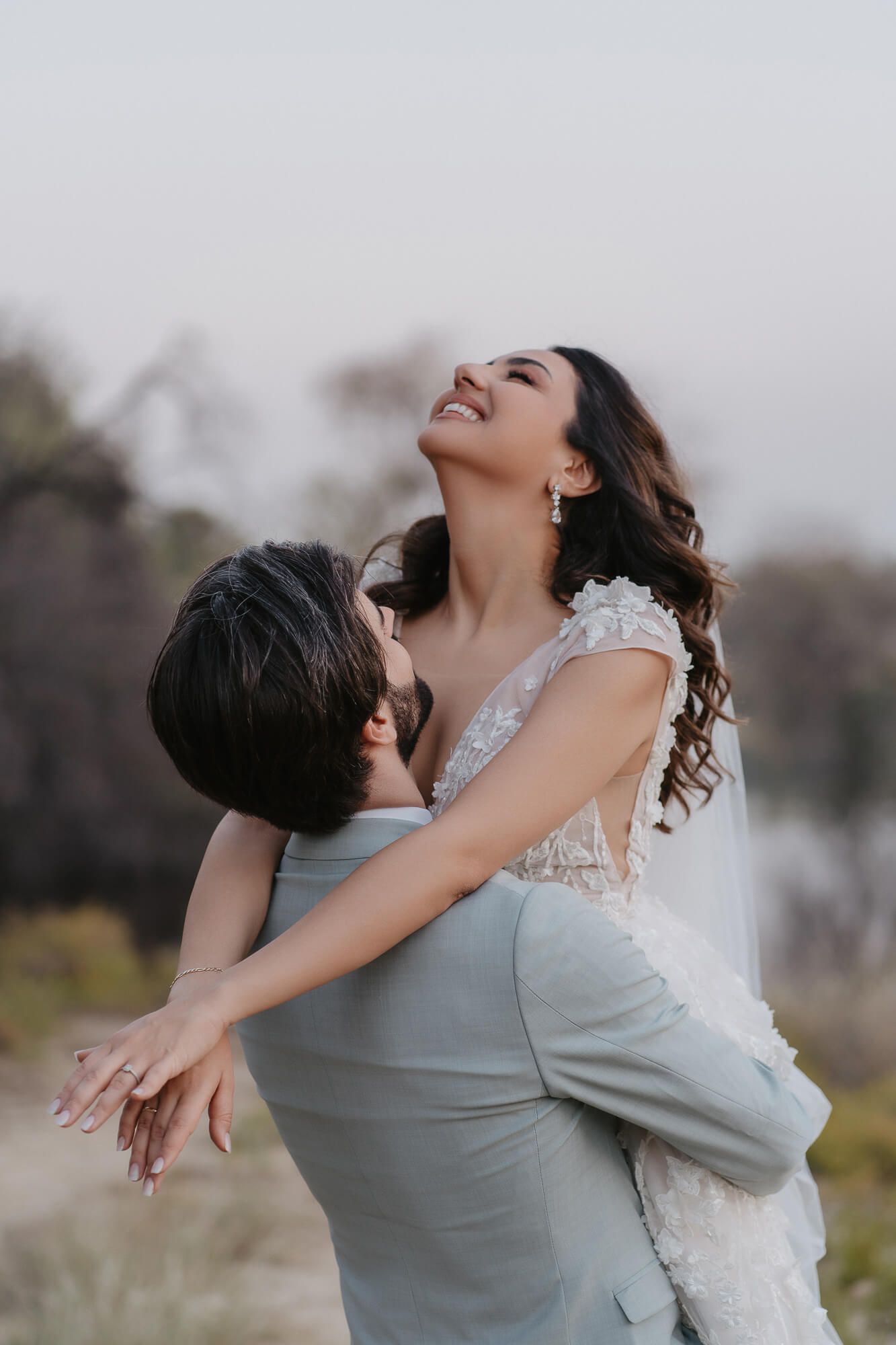 A couple embraces joyfully outdoors on their wedding day as the bride in white lace dress throws her head back laughing.