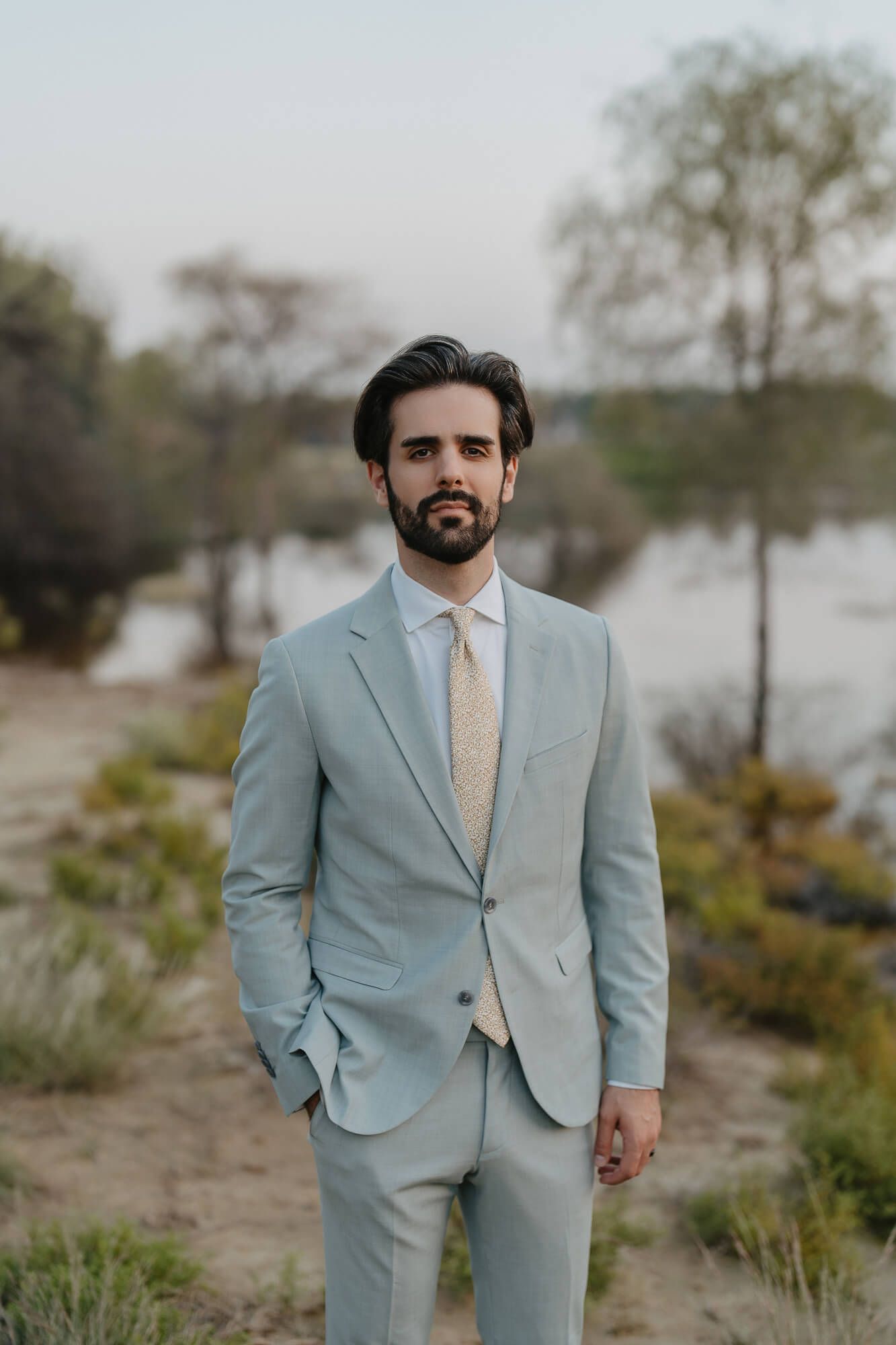 A person wearing a light blue suit and peach tie poses outdoors in a coastal setting for portraits.