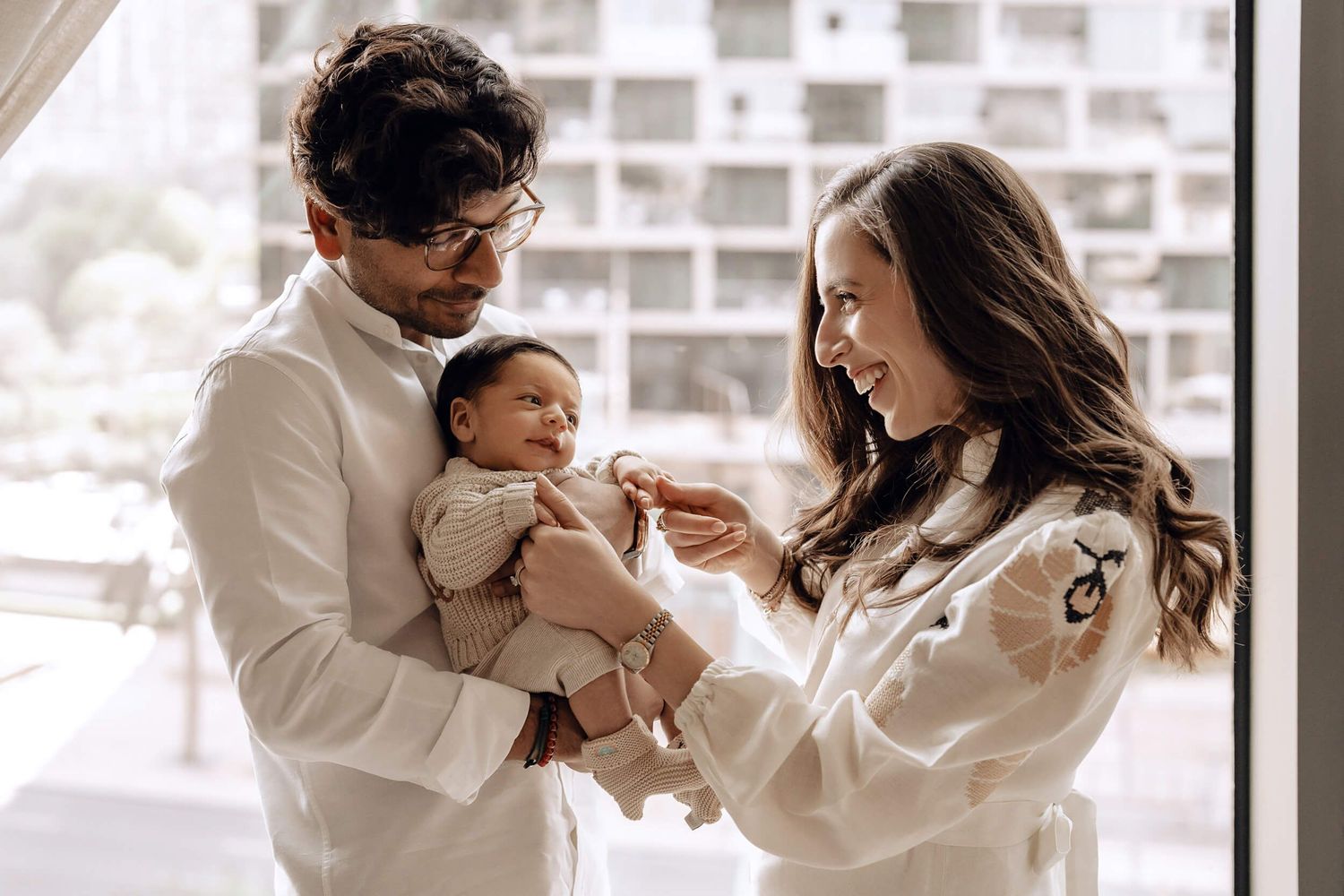A couple in white clothing hold and lovingly interact with their small baby near a bright window.