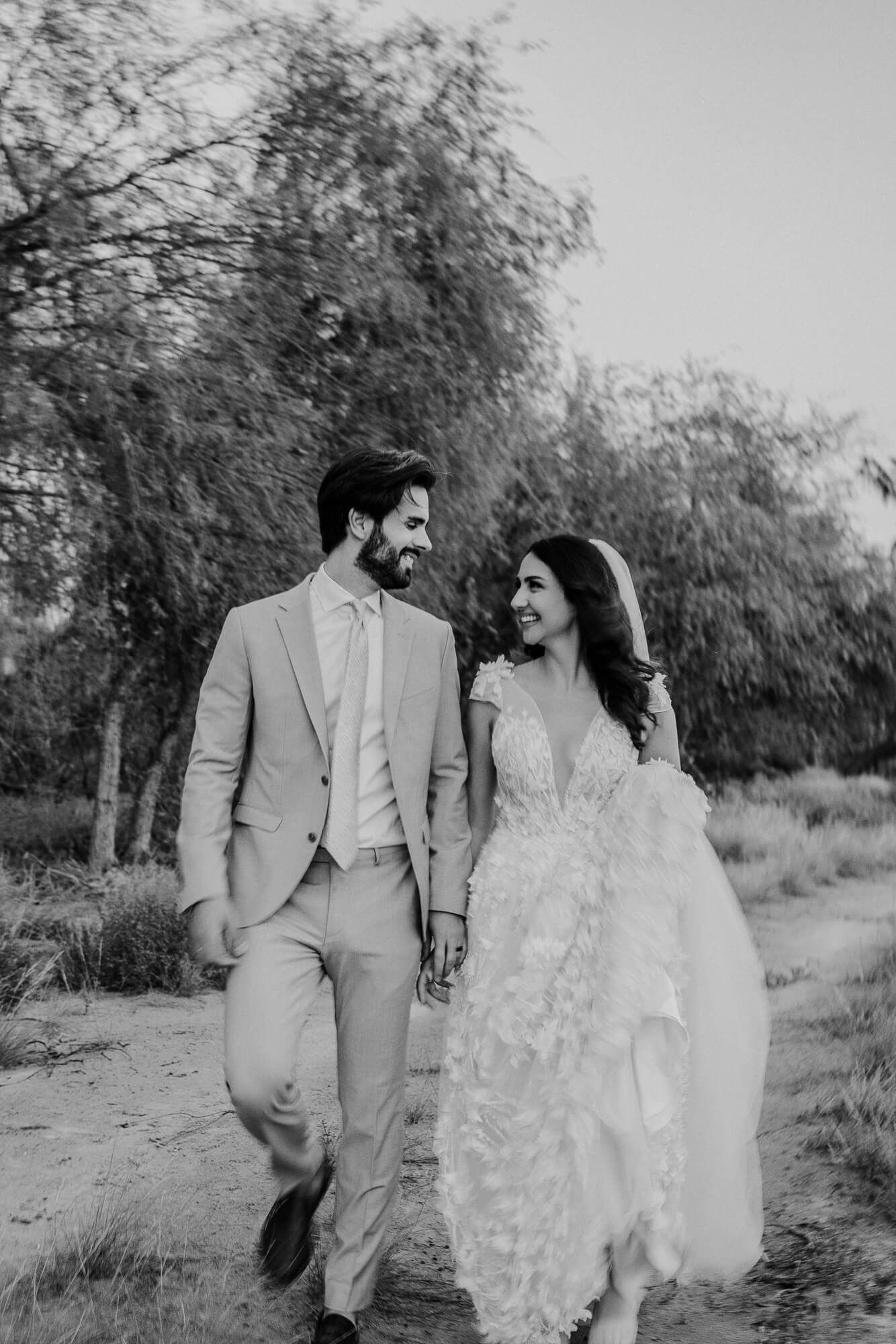 A couple in wedding attire walks together on a dirt path, with the bride in a flowing lace dress and groom in light suit.