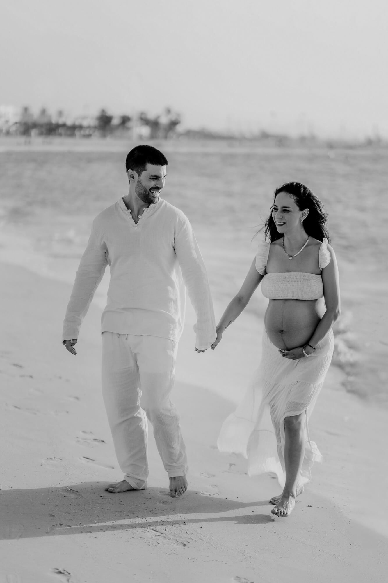 A black and white beach photo shows a couple in light colored clothing taking a romantic stroll in the sand.