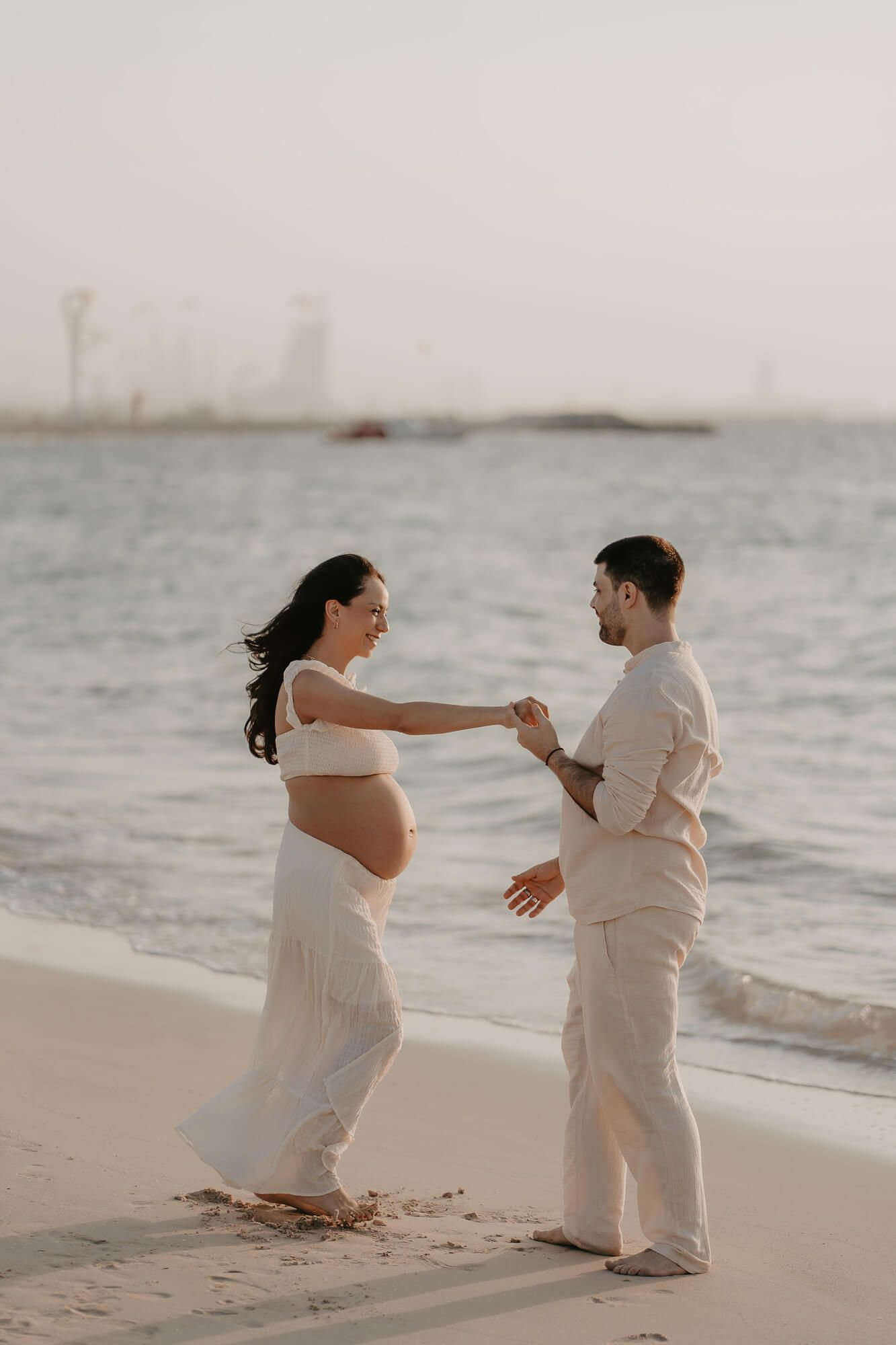 Couple in white outfits shares romantic moment on beach during sunset maternity photoshoot.