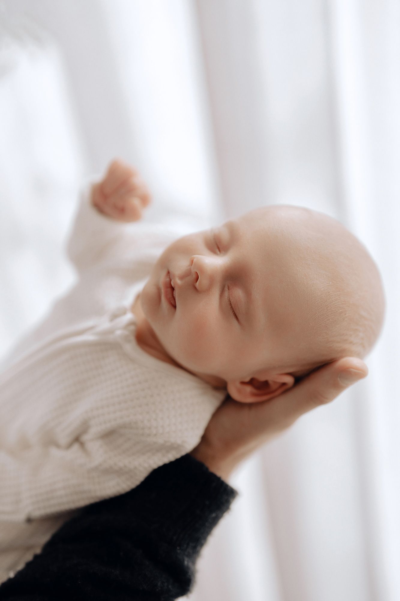 A peaceful sleeping newborn baby wearing a white knit sweater is cradled gently in someone's arms.