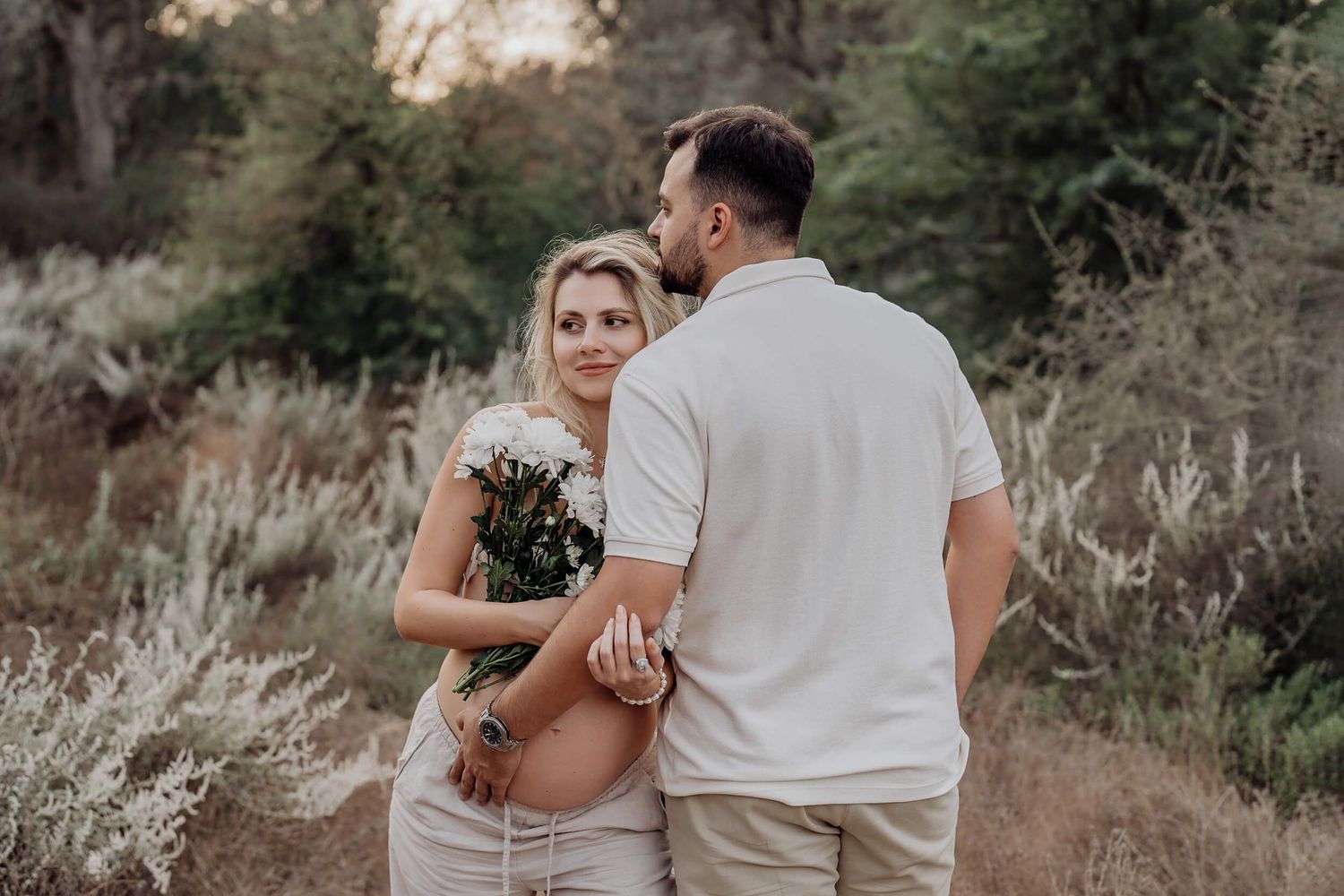 Candid maternity moment captured of couple in neutral tones standing together on outdoor path.