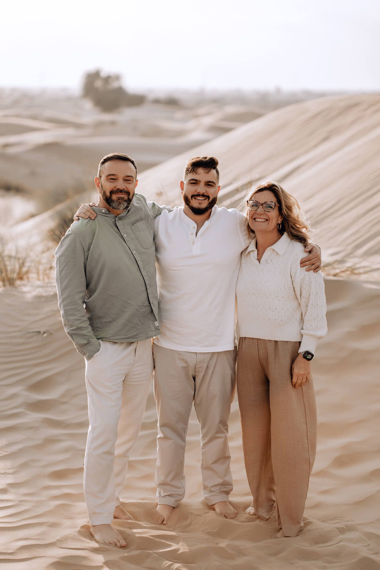 Three people wearing light colored clothing pose together on sandy desert dunes during sunset.