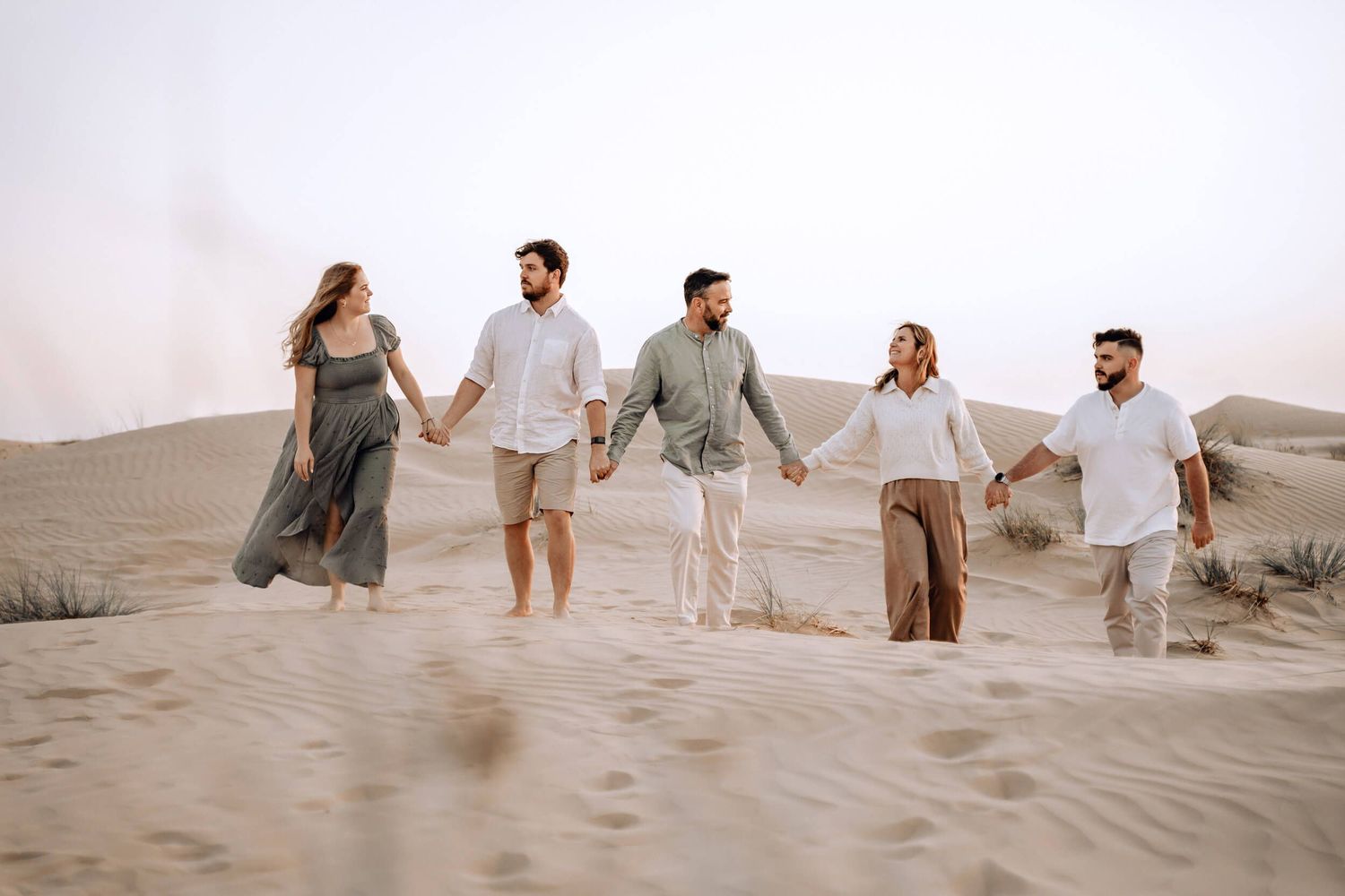 Group walking together and laughing on sandy desert dunes at sunset wearing casual neutral-toned clothing.